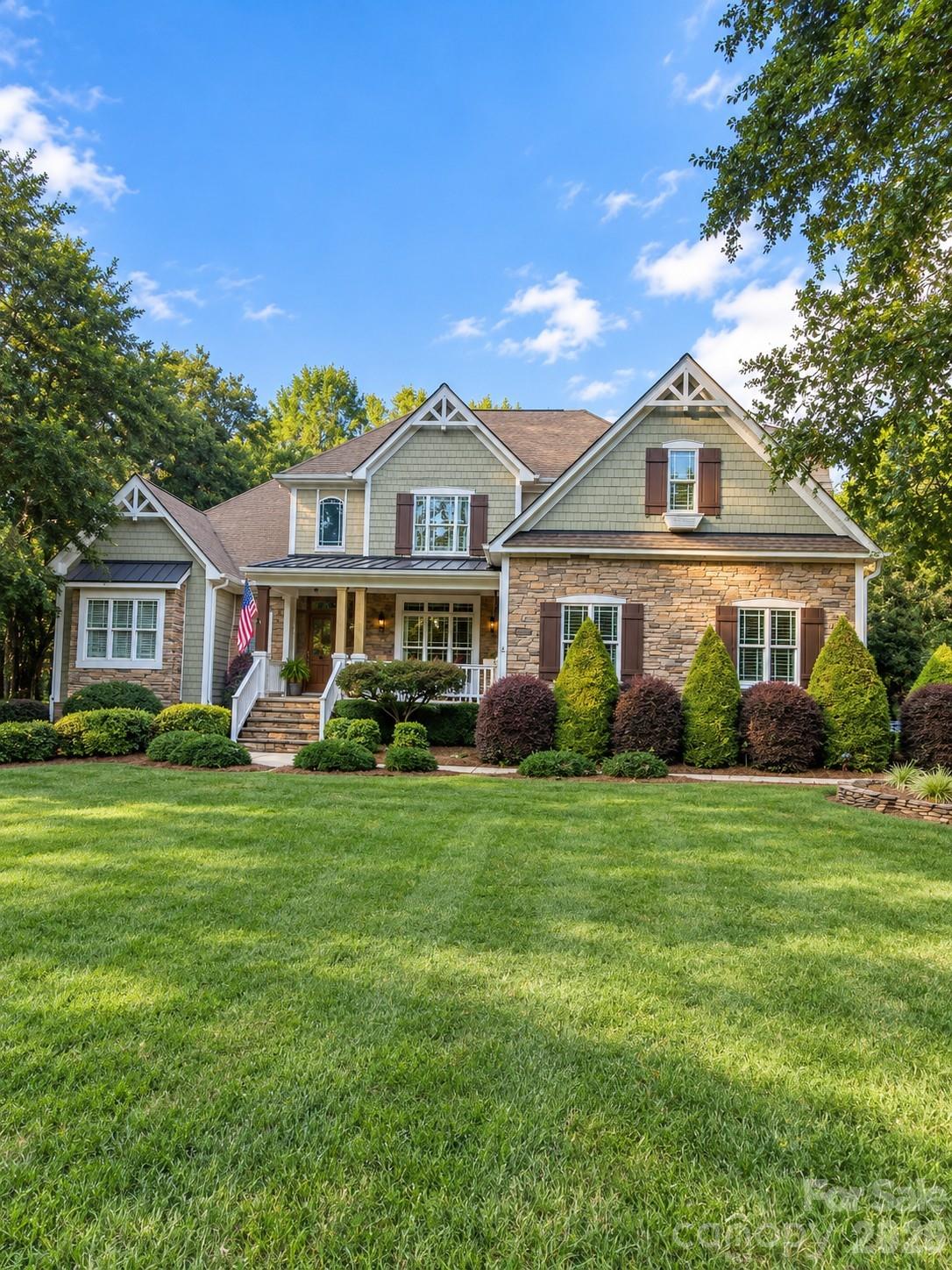 Two-story stone-and-shingle home with a covered porch, landscaped shrubs, and a wide green lawn.