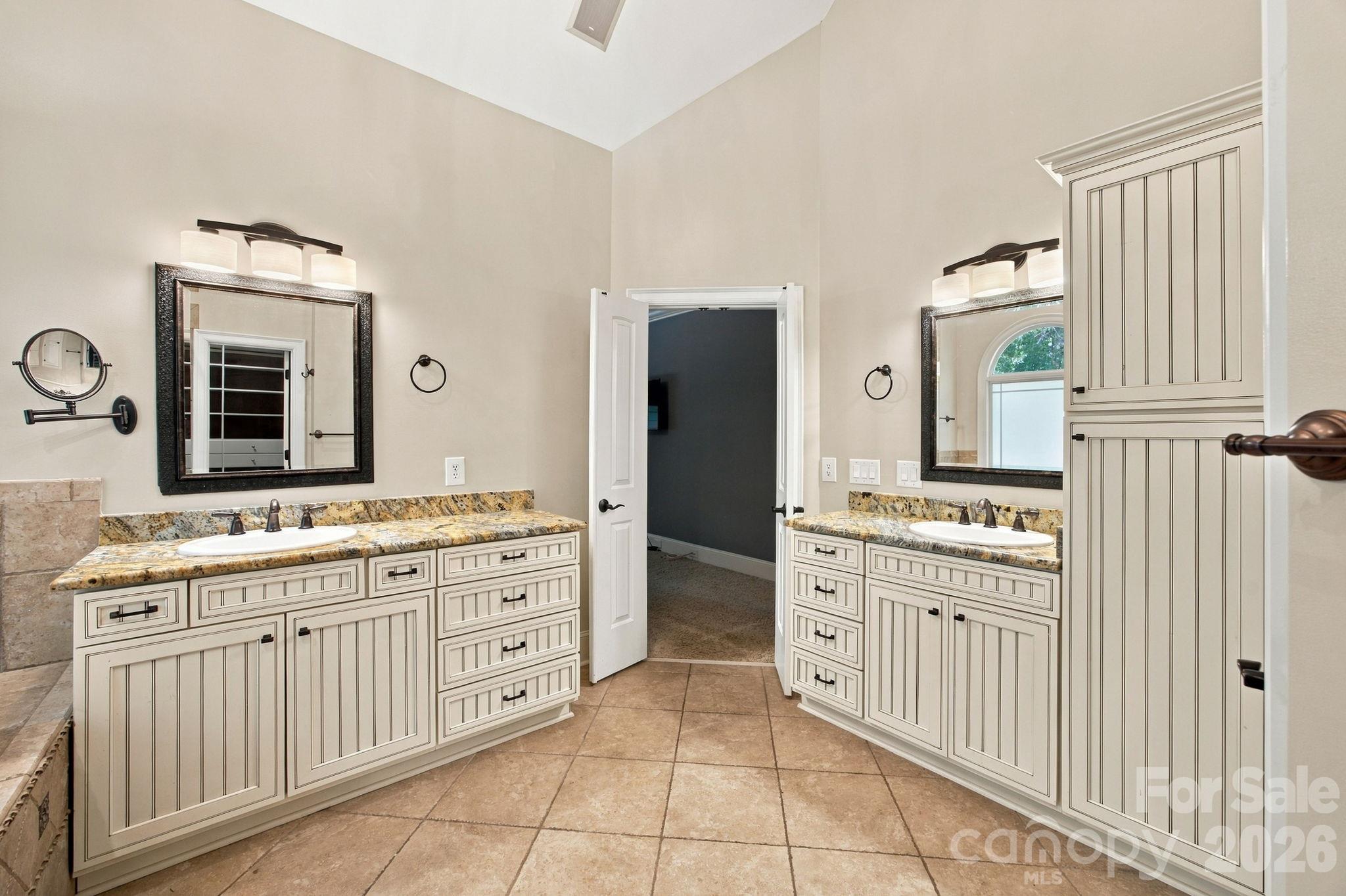 Bathroom with matching double vanities, granite countertops, large mirrors, and tan tile flooring.
