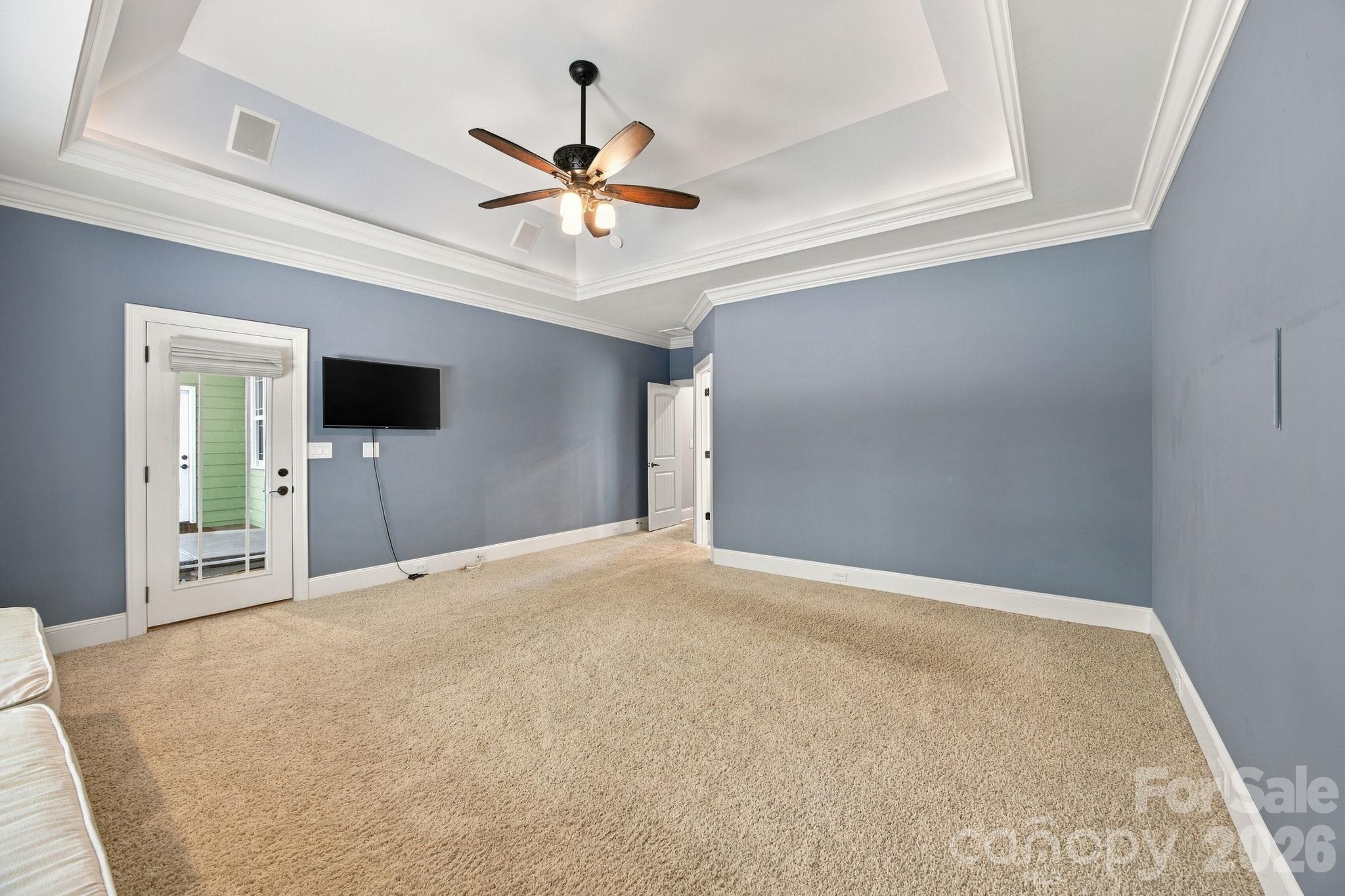 Bedroom with a tray ceiling, ceiling fan, beige carpet, and wall-mounted TV.