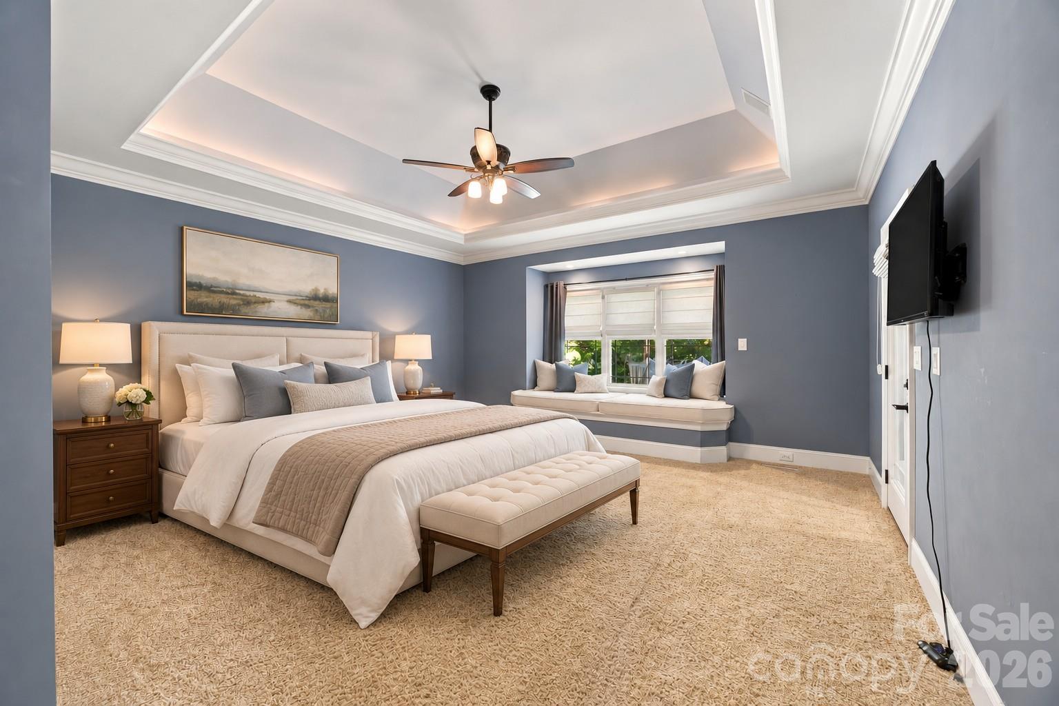Staged bedroom with a tray ceiling, upholstered bed, and window seat.