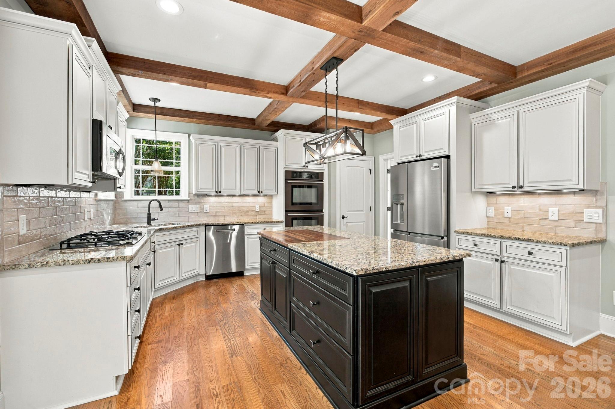 Kitchen with dark granite-topped island, white cabinetry, stainless steel appliances, and exposed wood ceiling beams.