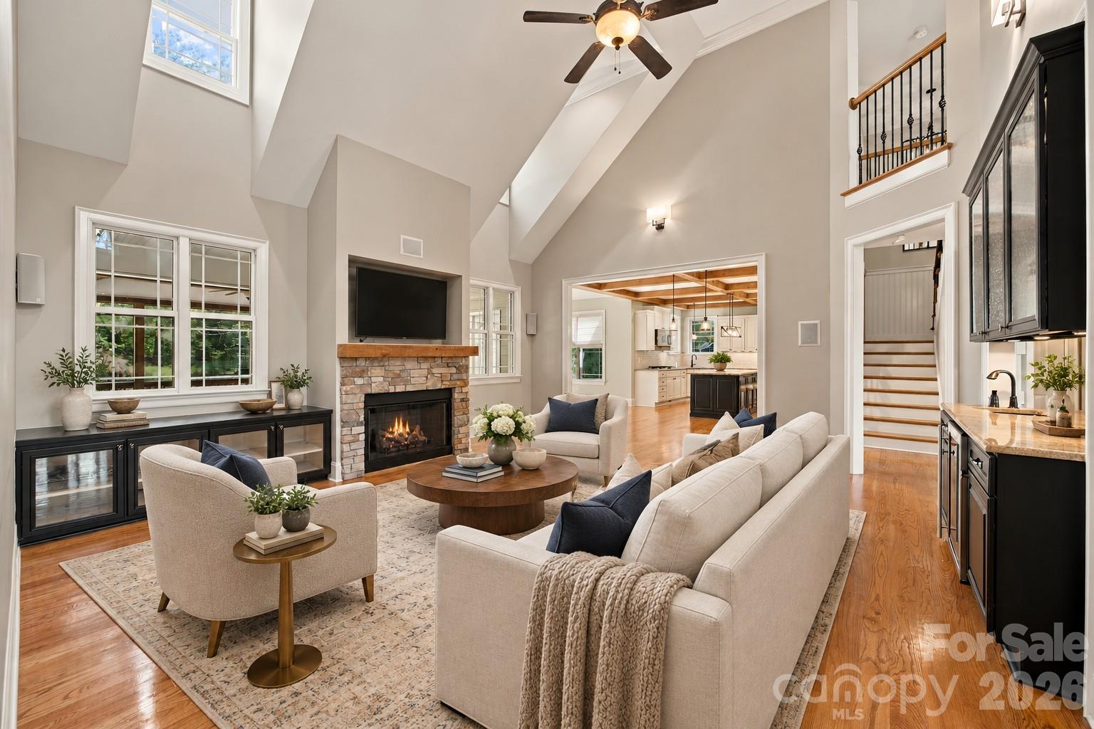 Vaulted living room with a stone fireplace, large windows, and an open view into the kitchen.