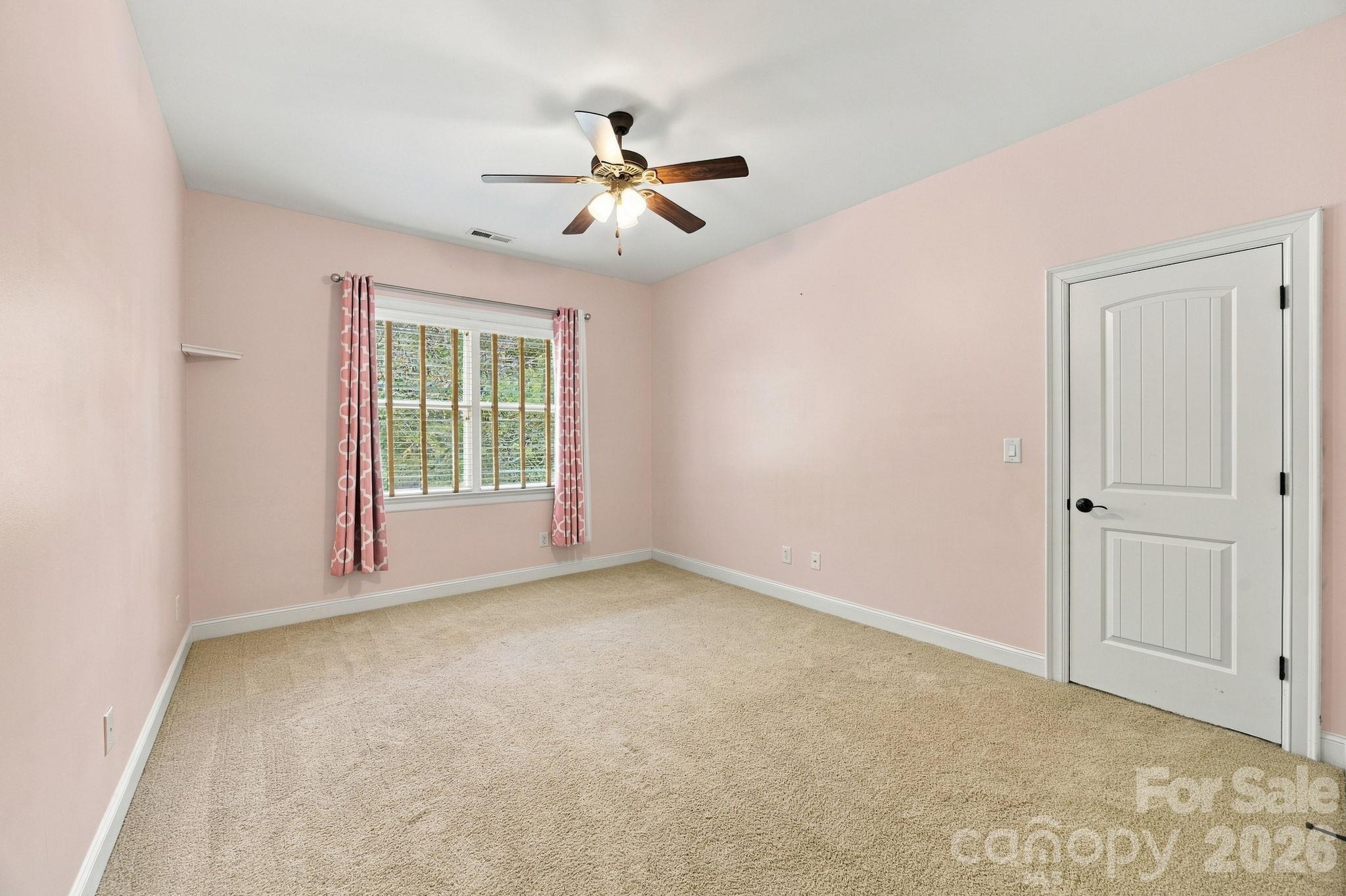 Empty bedroom with pale pink walls, beige carpet, pink curtains, and a ceiling fan.