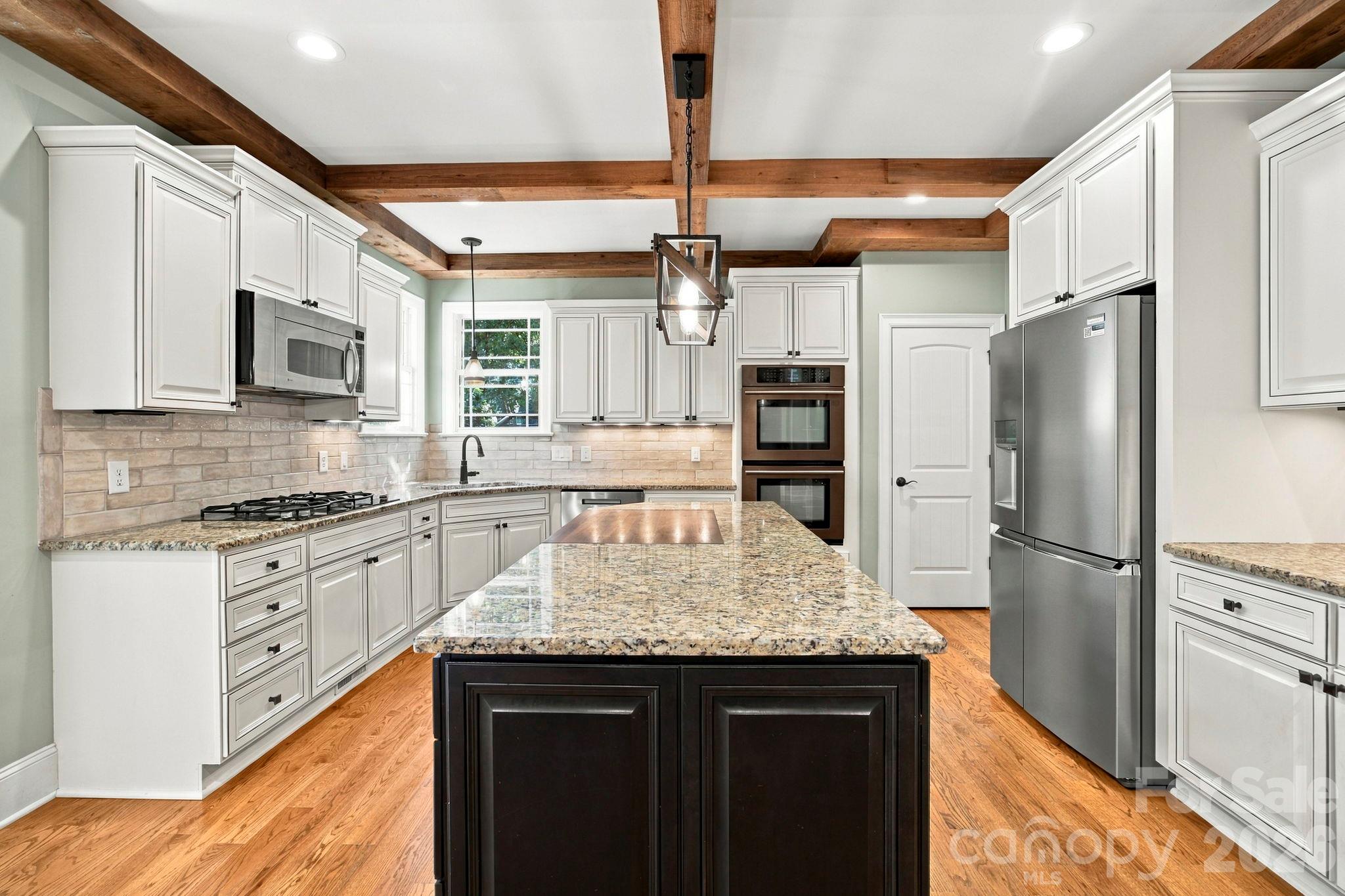Kitchen with a large granite island, white cabinets, stainless steel appliances, and exposed wood ceiling beams.