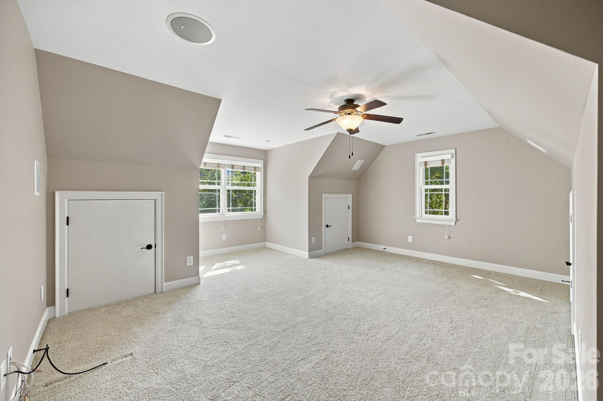Empty carpeted upper-level room with sloped ceilings, two windows, and a ceiling fan.