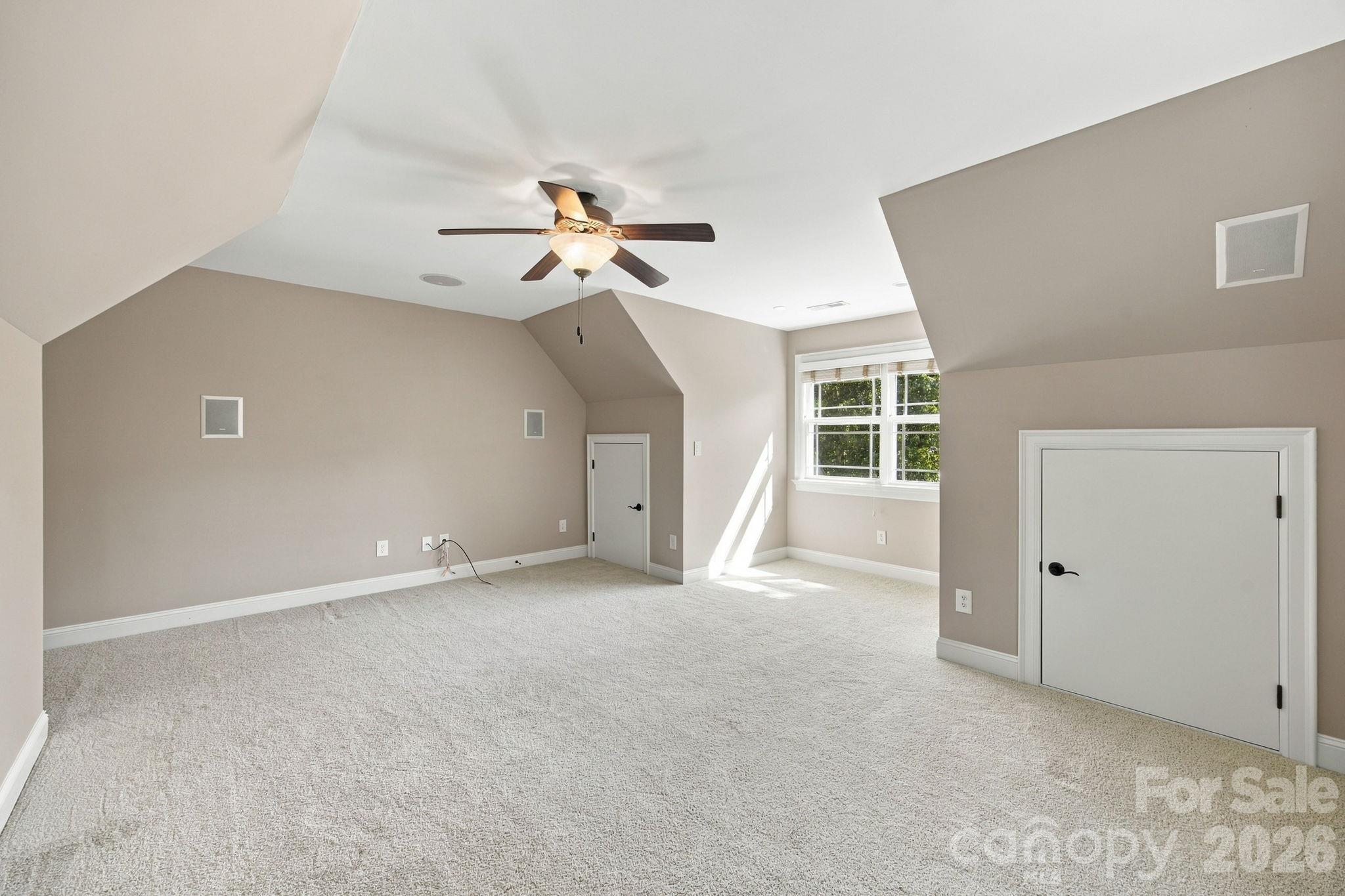 Empty carpeted room with a ceiling fan, angled ceiling, and sunlit window.