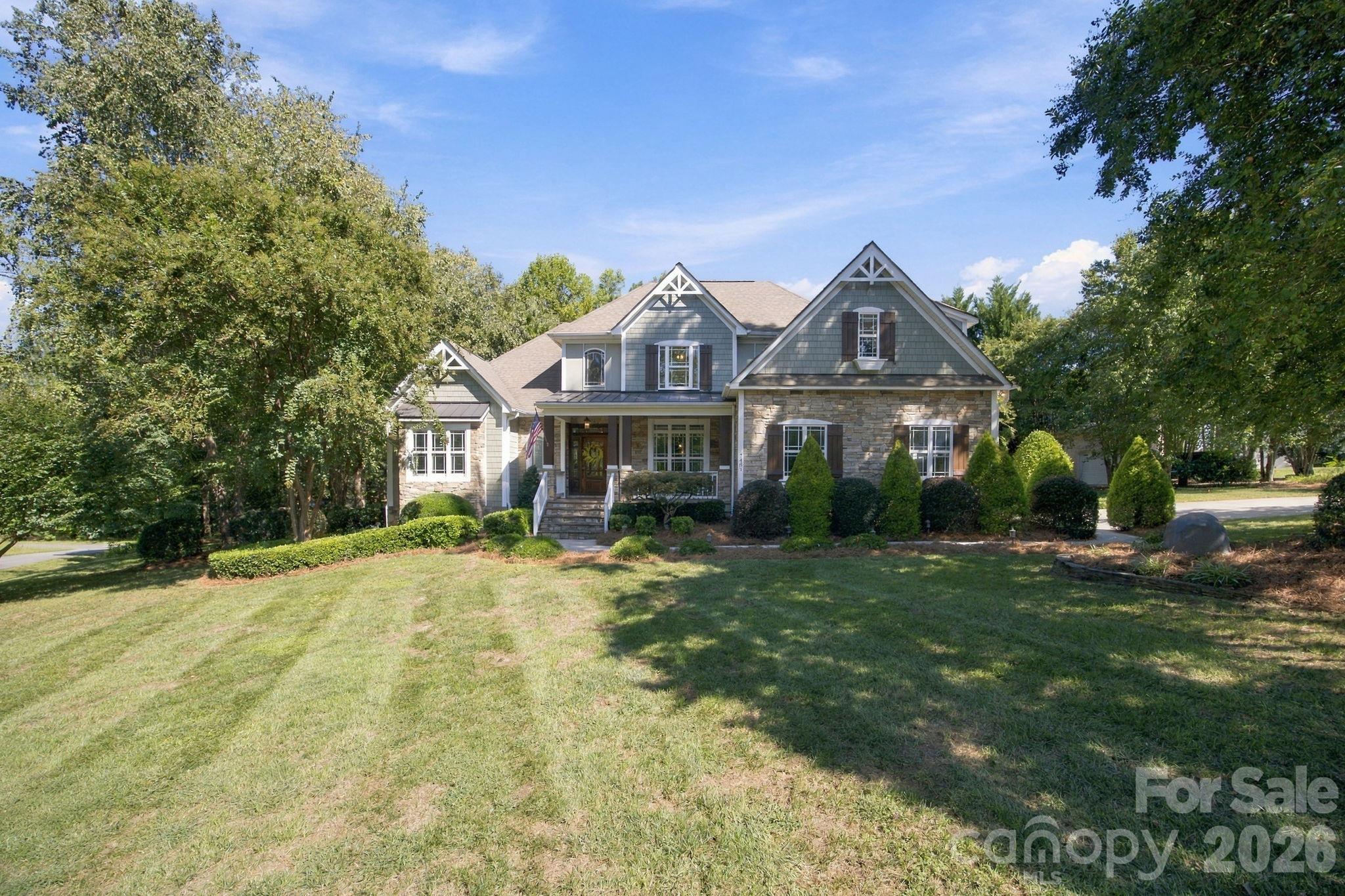 Two-story gray-shingled and stone house with a covered porch, mature trees, and a broad front lawn.
