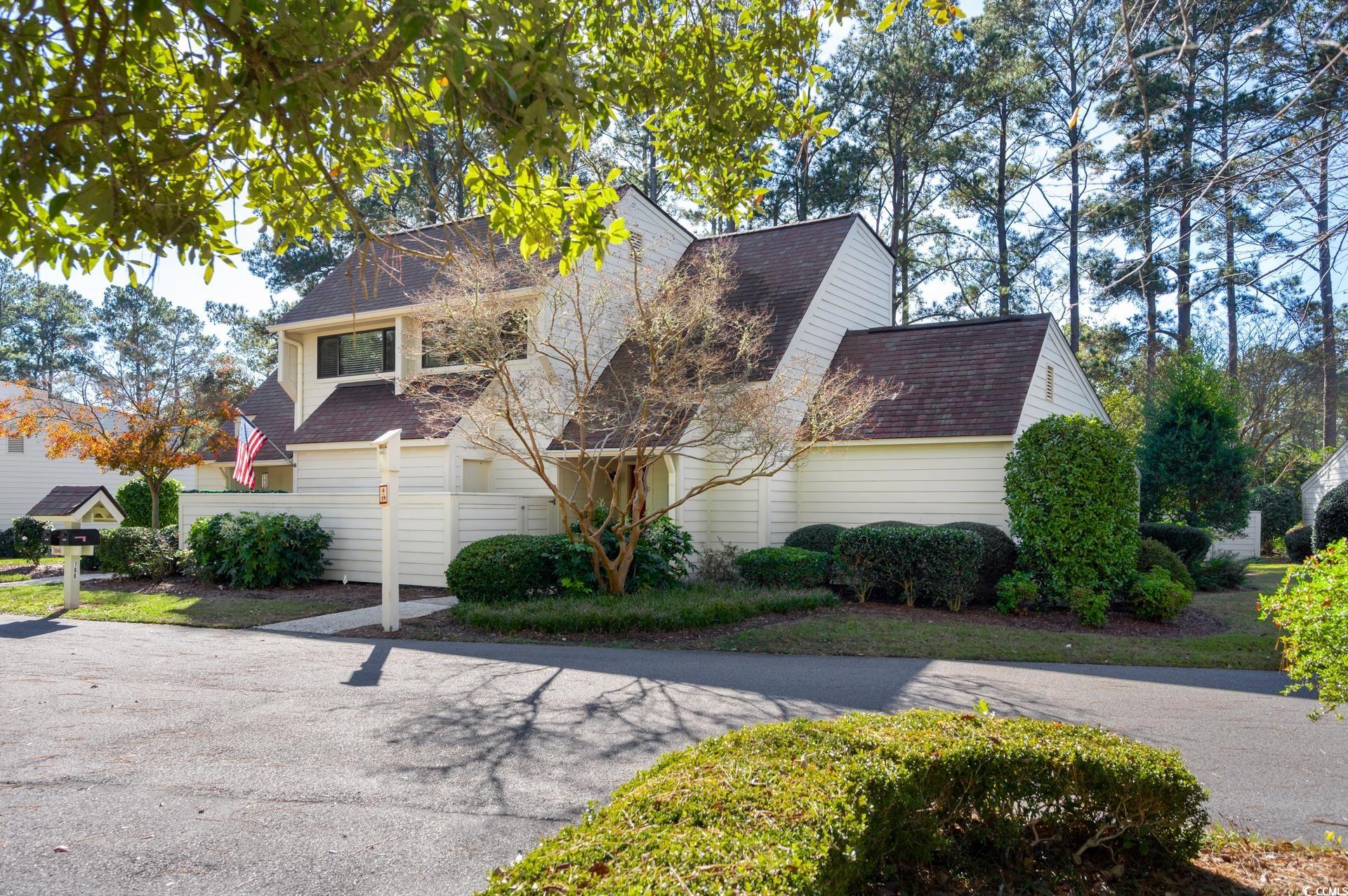 White two-story home with dark gabled roofs, a privacy fence, and an American flag among mature trees.