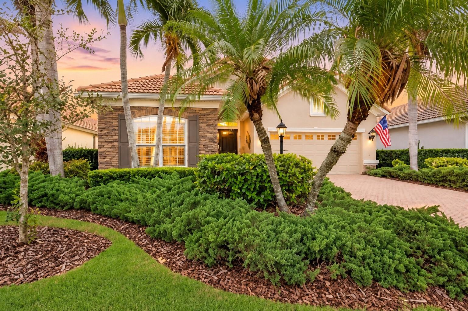 Stucco home with a stone facade, terracotta roof, palm-lined landscaping, and an American flag by the garage.
