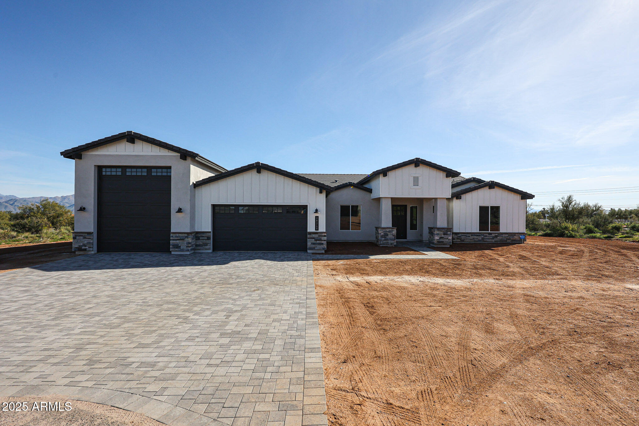 Modern white-and-gray home with multiple dark garage doors, a paved driveway, and an unfinished dirt yard.