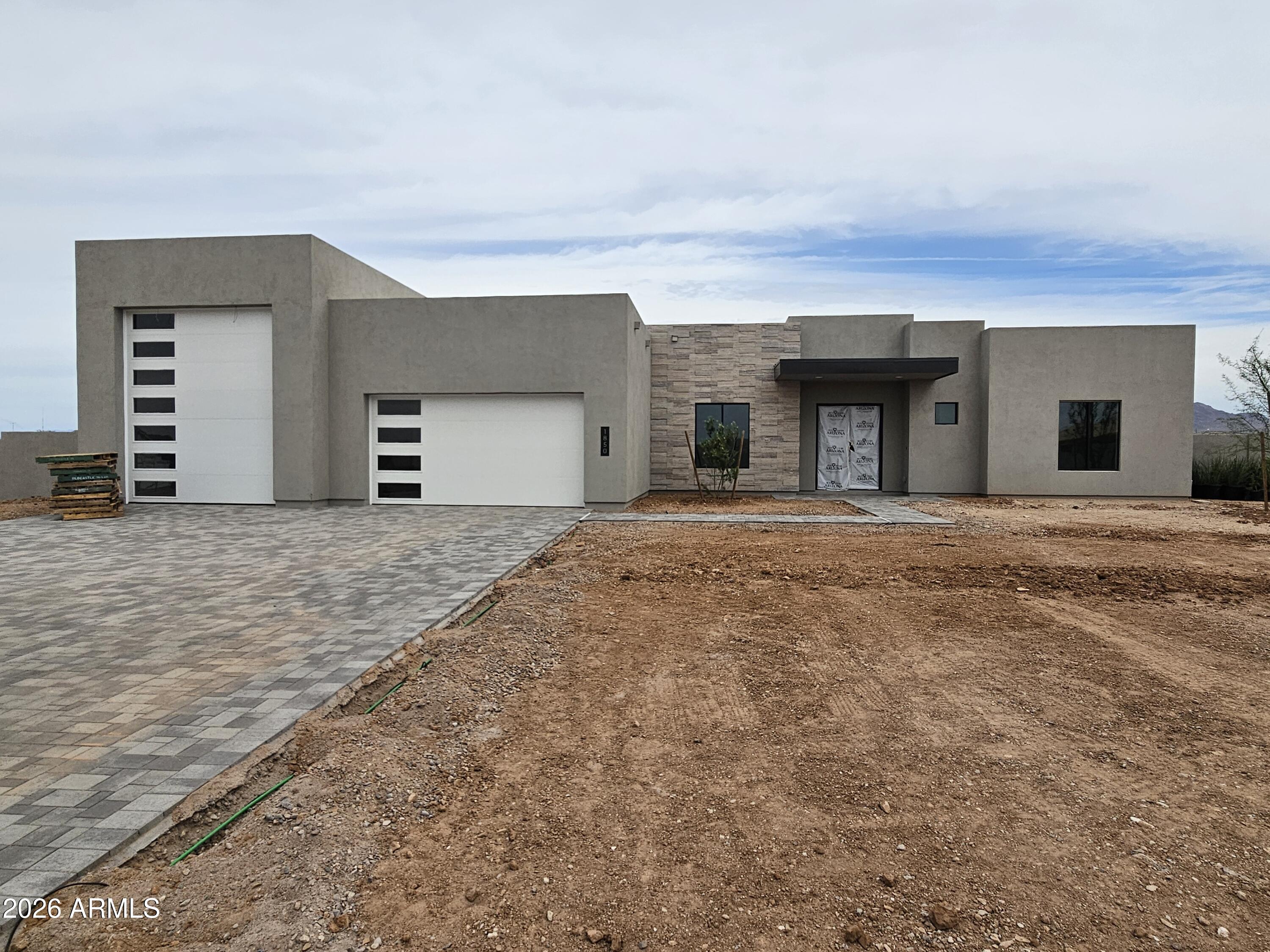 Modern gray stucco home with two white garage doors, stone entry, and an unfinished dirt front yard.