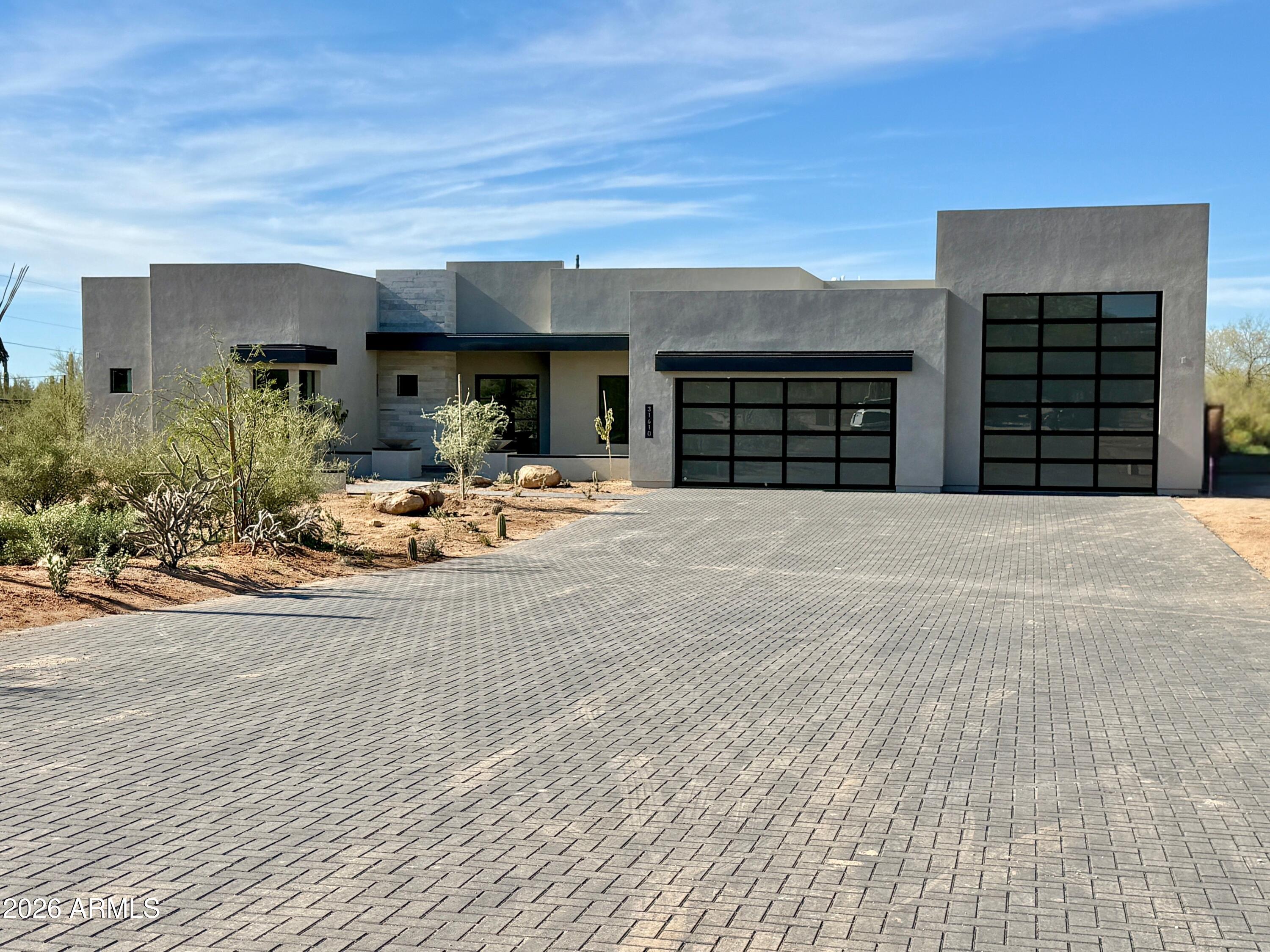Modern gray home with two black-framed garage doors, a broad paver driveway, and desert landscaping.