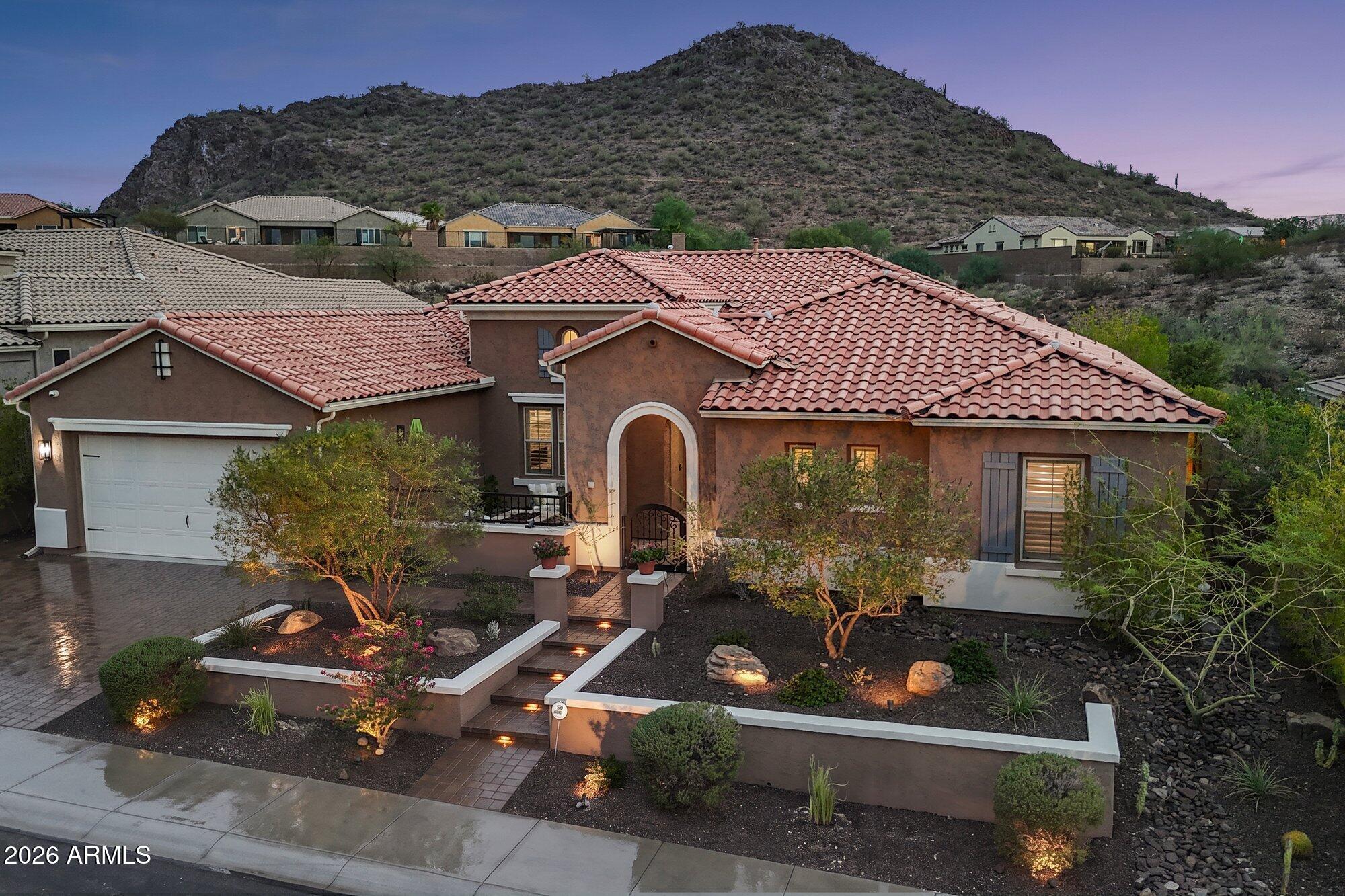 Stucco home with red tile roofs, an arched entry, desert landscaping, and a mountain backdrop.