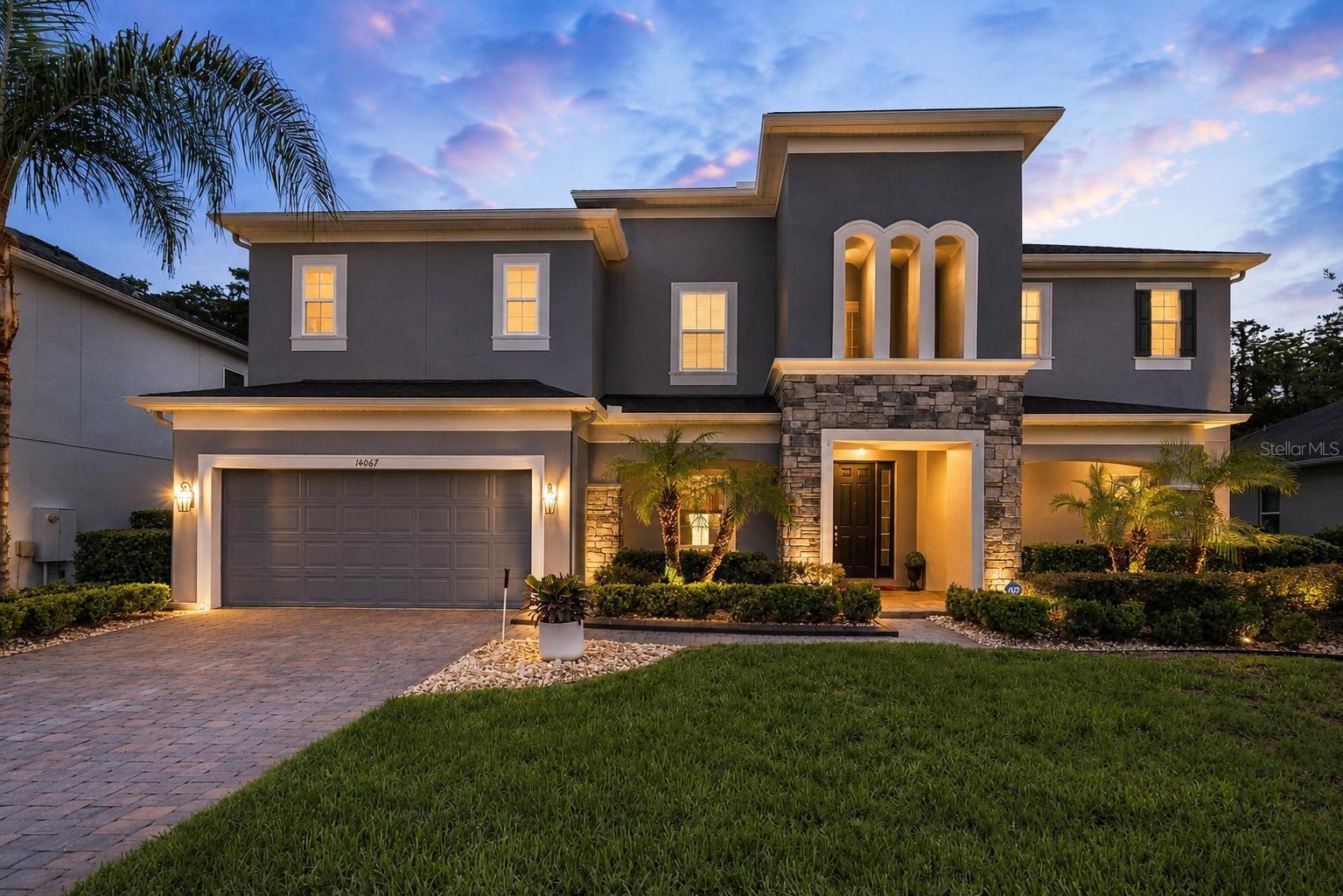 Two-story gray house with a stone entry, arched windows, double garage, and landscaped front yard at dusk.