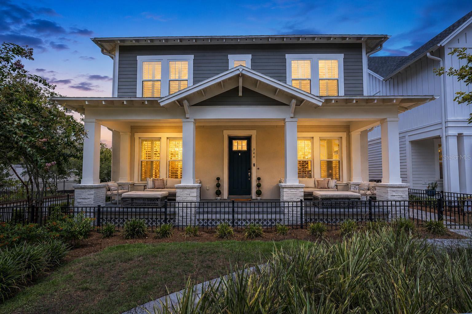 Two-story gray house with a furnished covered porch and glowing windows at dusk.