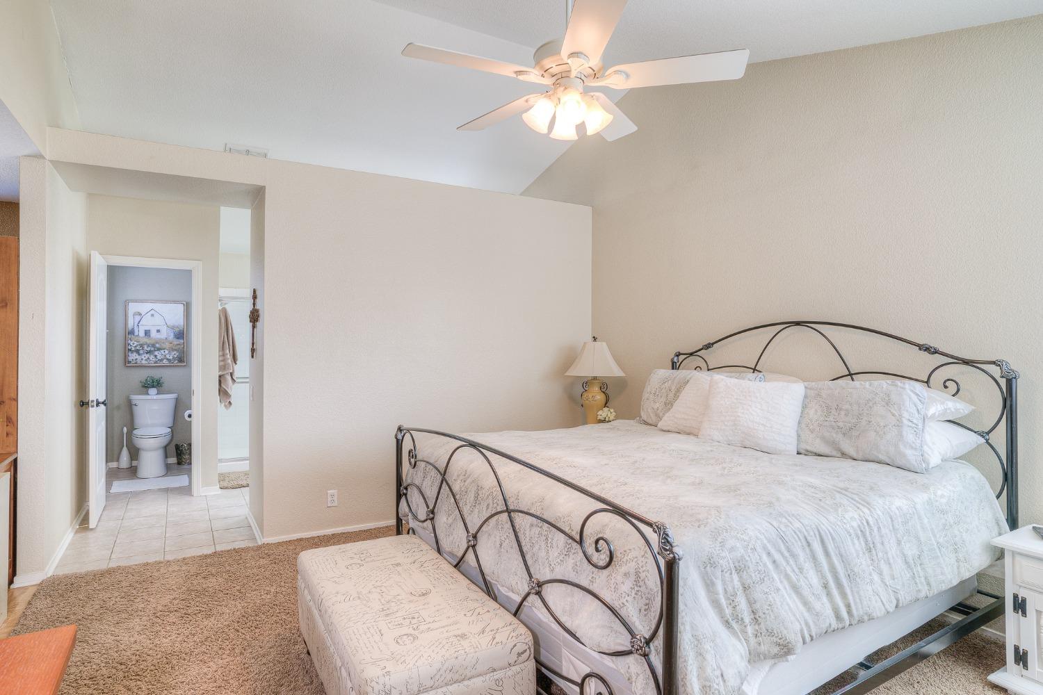 Furnished bedroom with a metal-framed bed, upholstered bench, ceiling fan, and open bathroom doorway.