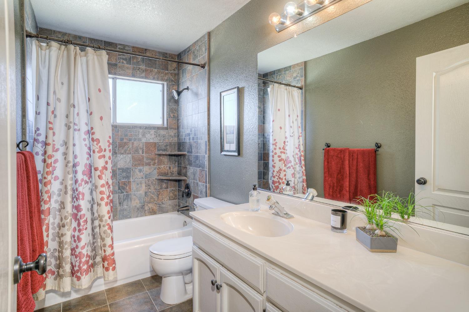 Bathroom with tiled tub and shower, patterned curtain, and long white vanity with a large mirror.