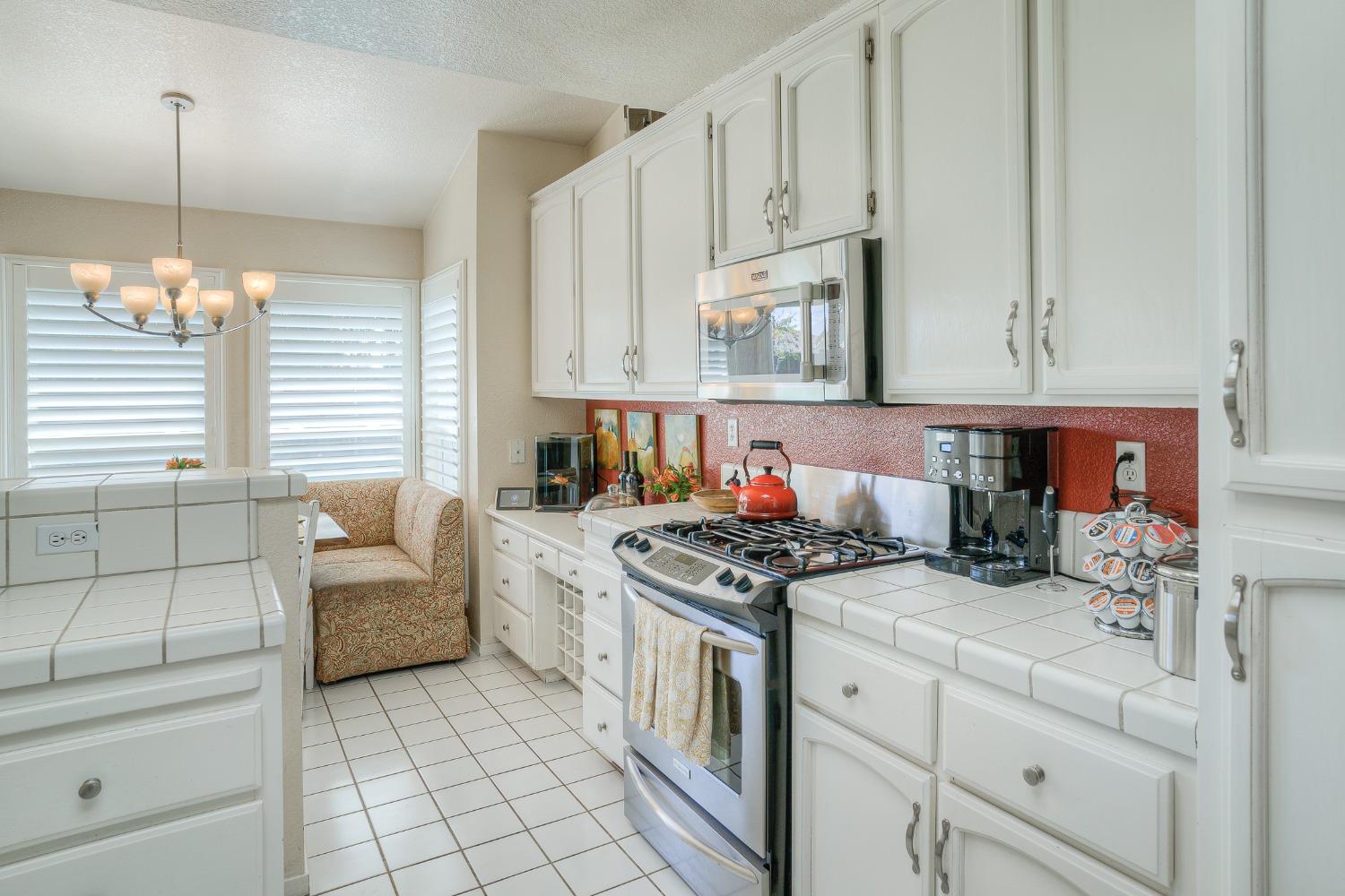 Bright kitchen with white cabinets, a stainless-steel gas range, tiled counters, and a shuttered breakfast nook.