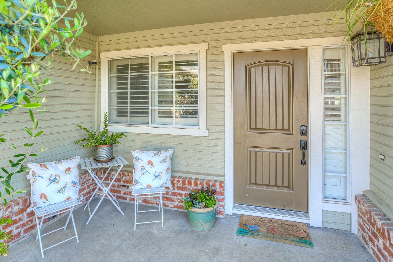 Covered front porch with a tan entry door, white chairs, bird-print cushions, and potted plants.