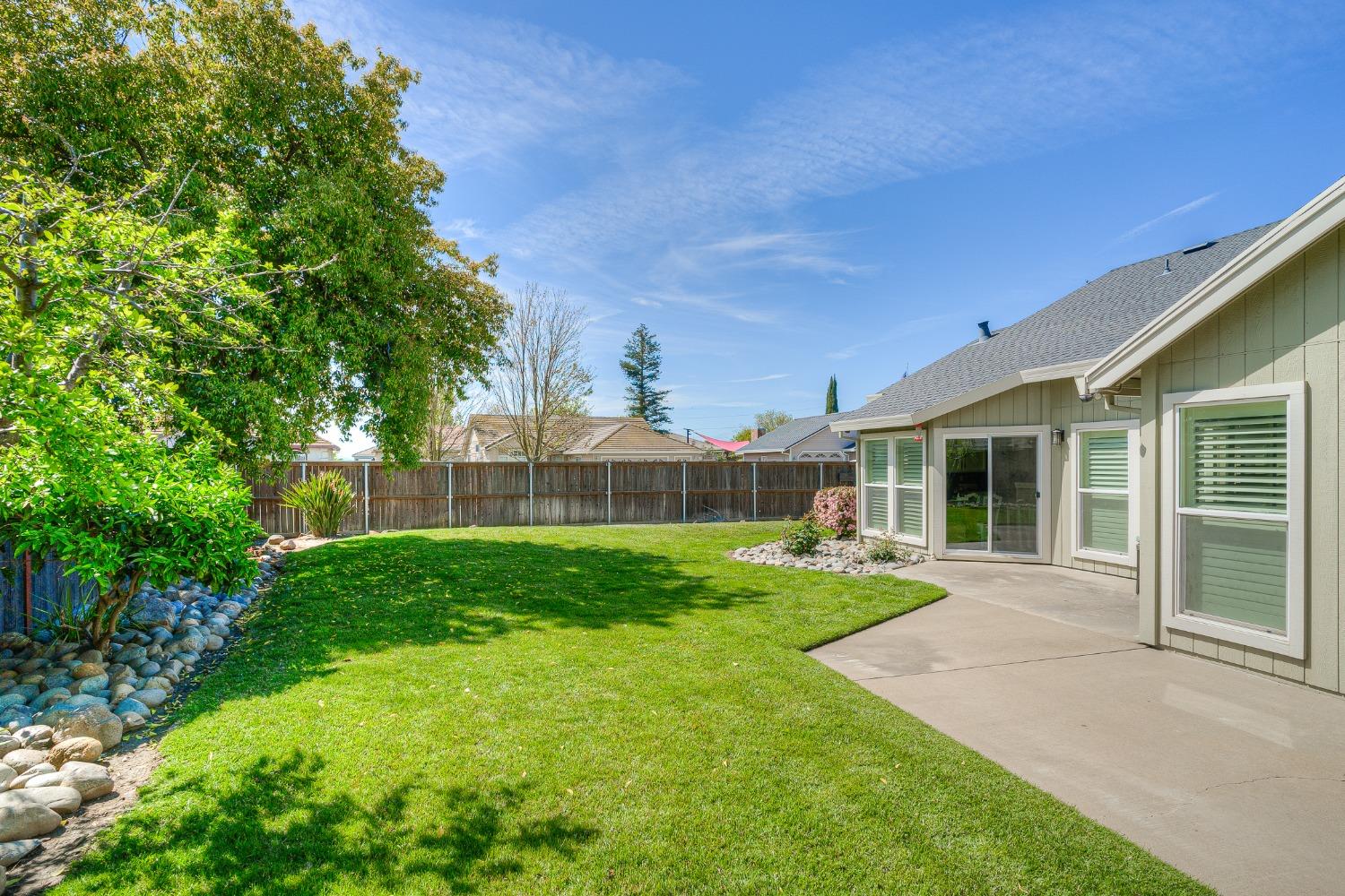 Fenced backyard with a broad lawn, stone-edged landscaping, mature trees, and a concrete patio.