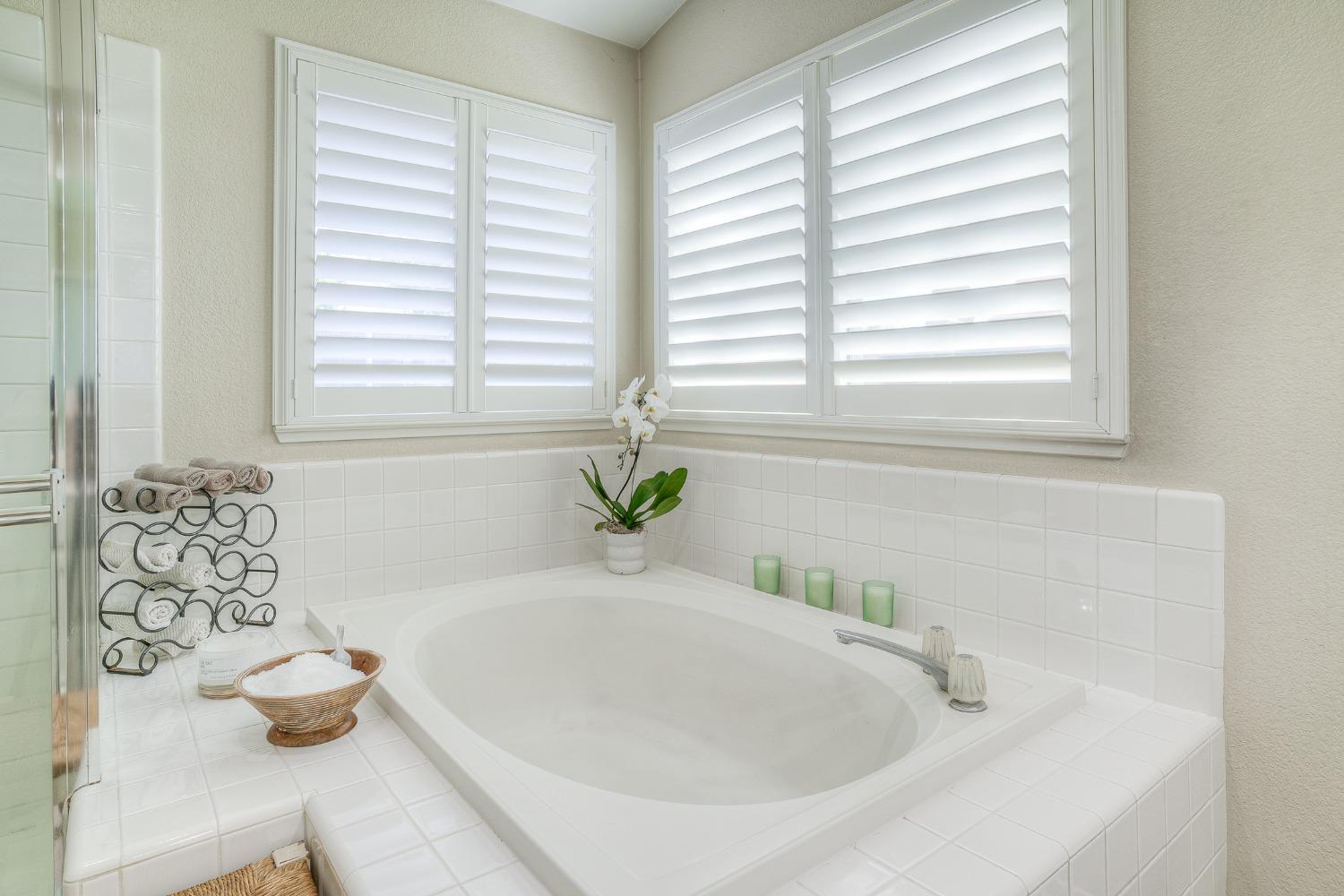 Bathroom soaking tub beneath corner windows with white plantation shutters and tiled surround.