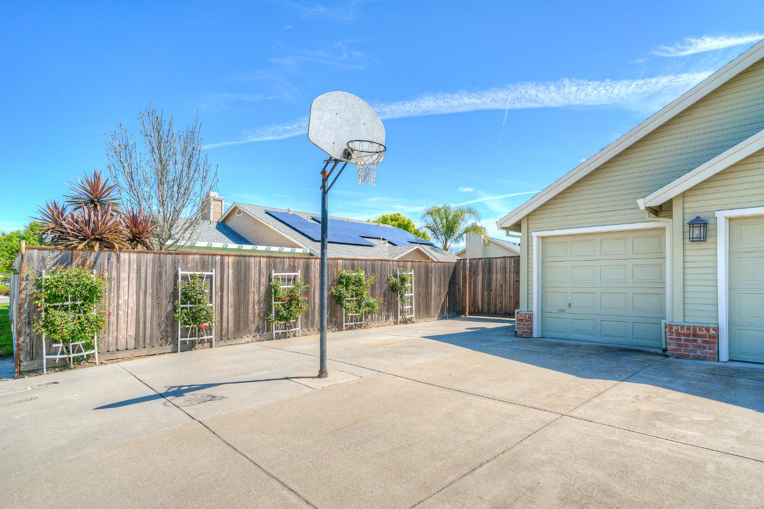 Concrete driveway beside a garage, with a basketball hoop and fenced rose garden.