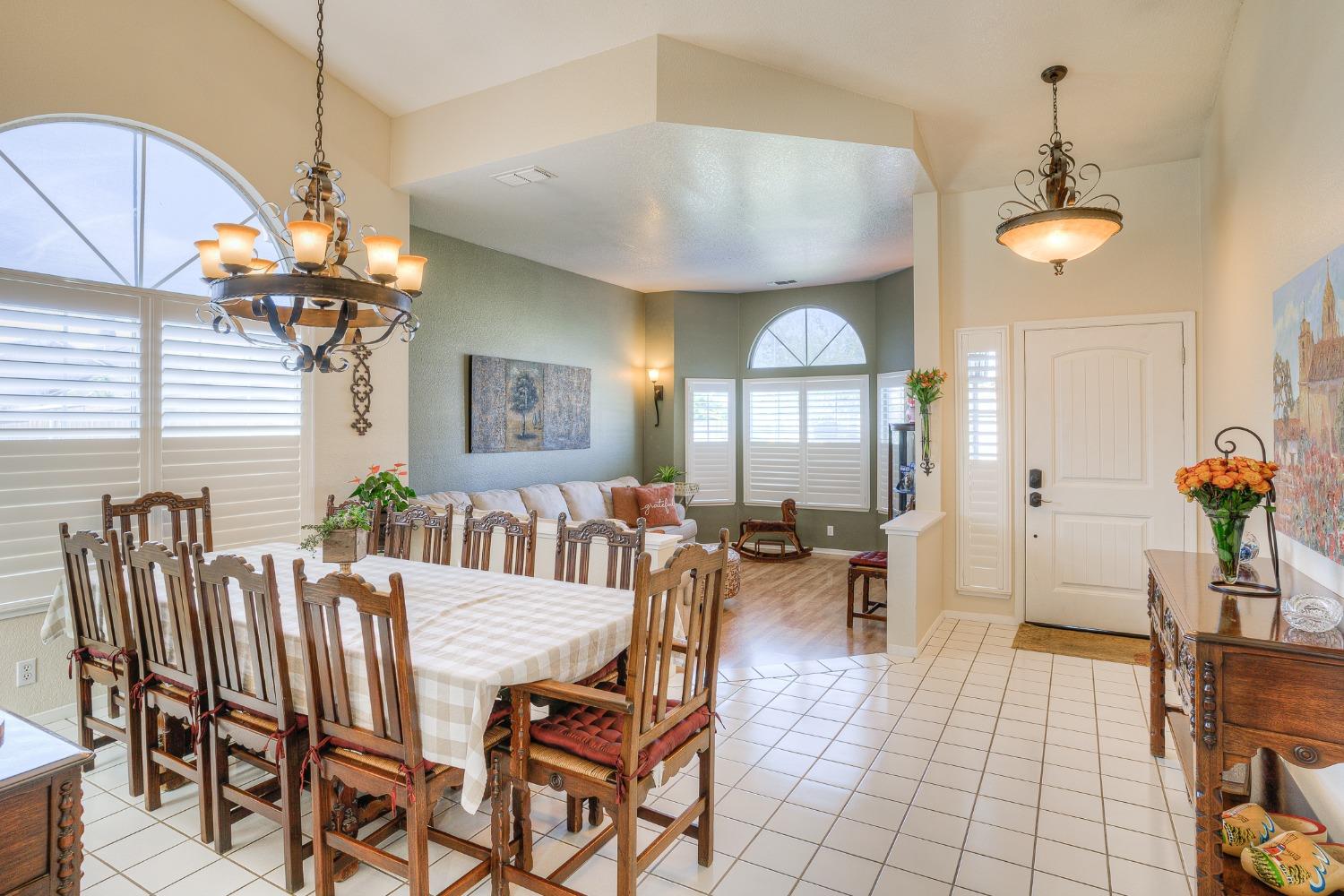 Dining room with a large wooden table, ornate chandelier, plantation shutters, and adjoining living area.