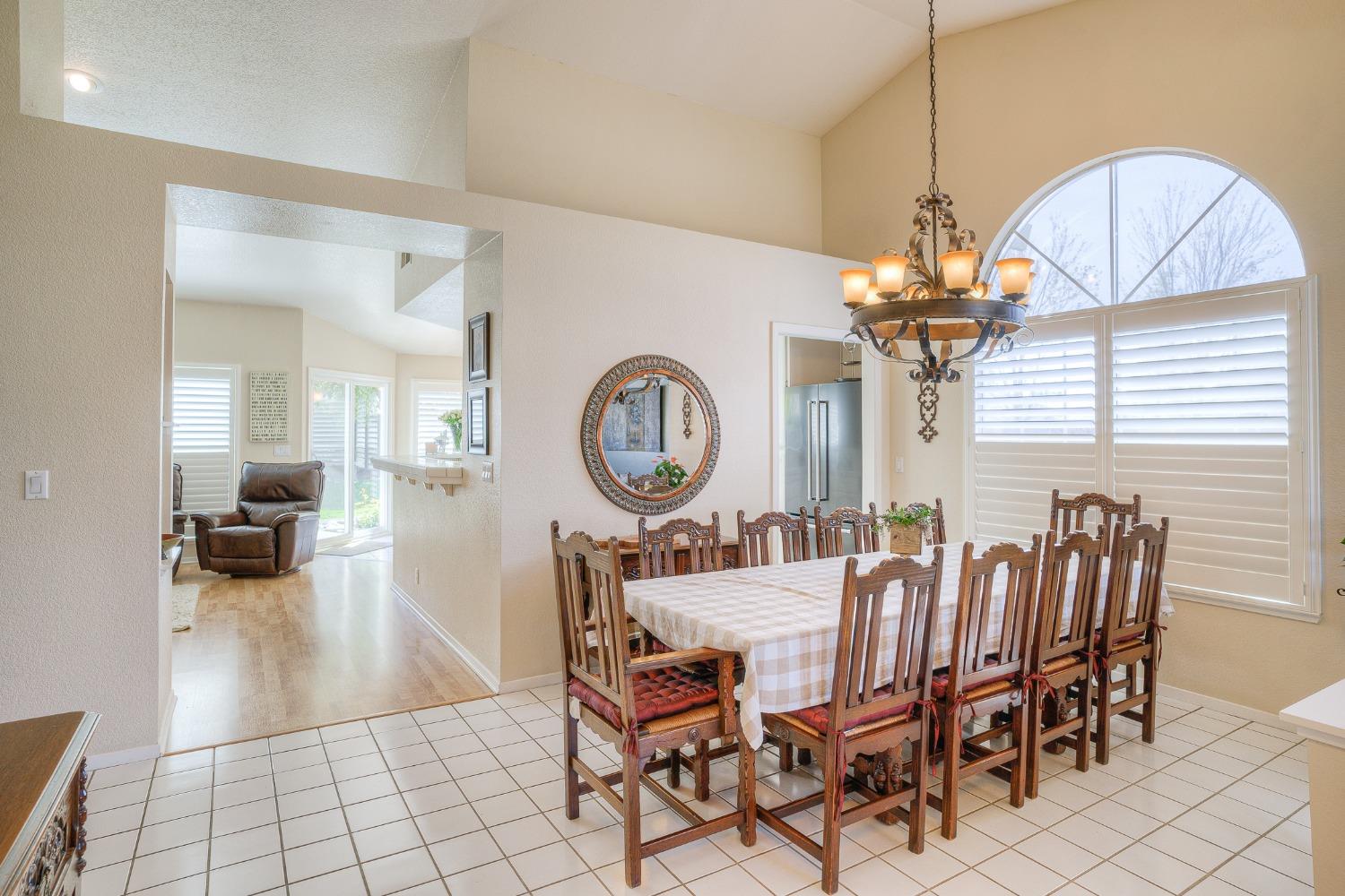Dining room with a large wooden table, ornate chandelier, and arched window with plantation shutters.