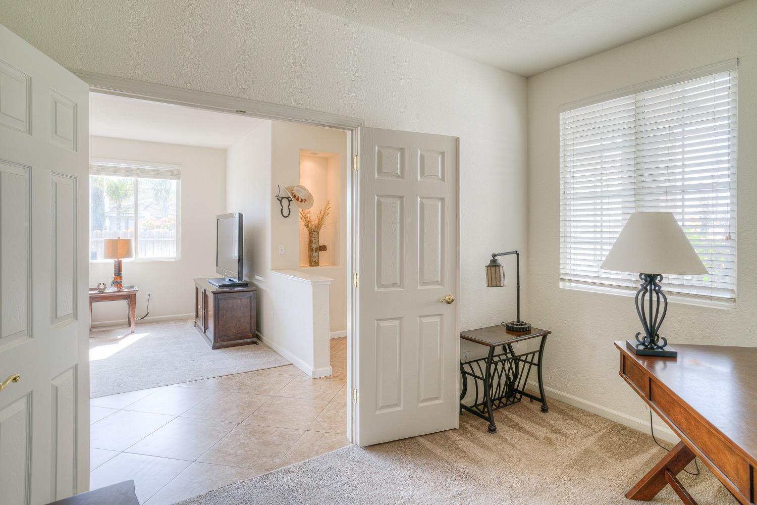Carpeted sitting room with a wooden desk and lamp beside an open doorway to a tiled room with a television.