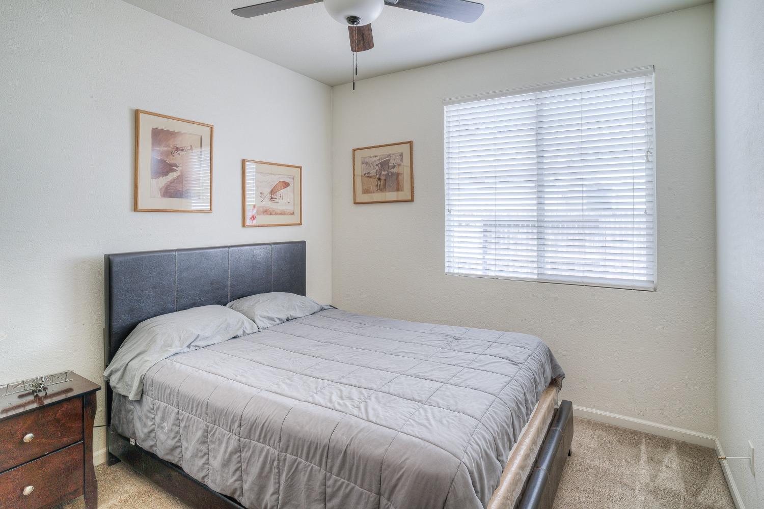 Bedroom with a gray quilted bed, dark headboard, white blinds, and framed wall art.