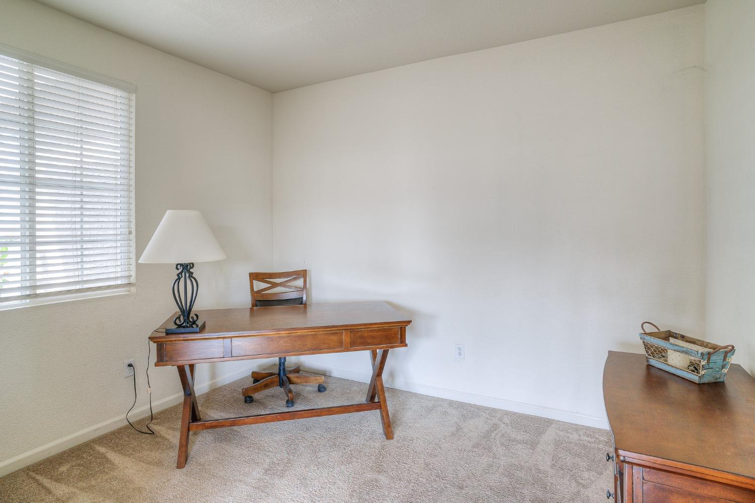 Carpeted home office with a wooden desk and chair, dresser, and window with white blinds.