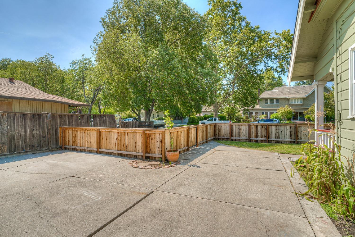 Concrete backyard patio enclosed by wooden fencing, with mature trees beyond.