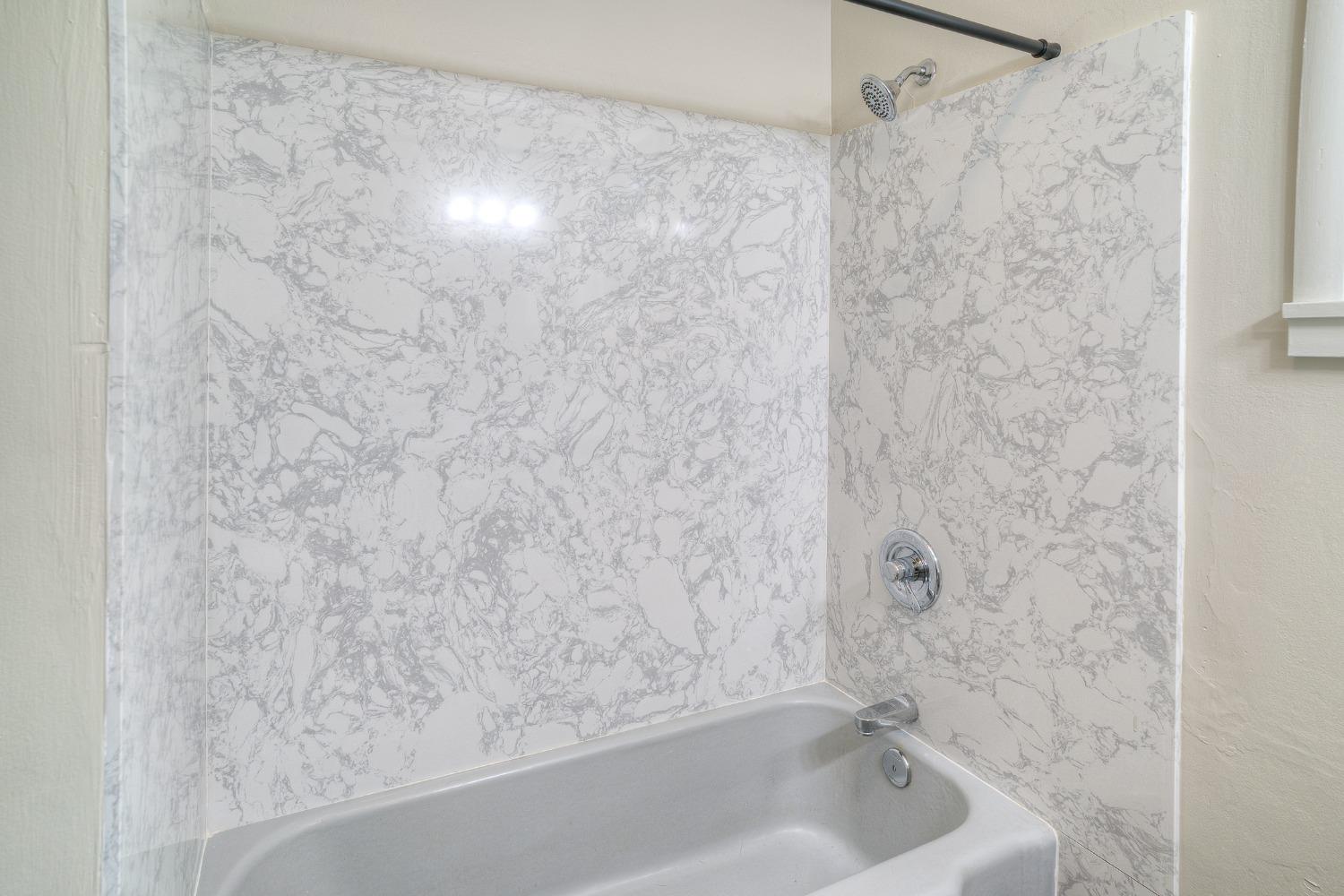 Bathroom bathtub with a gray-veined marble-pattern shower surround and chrome fixtures.