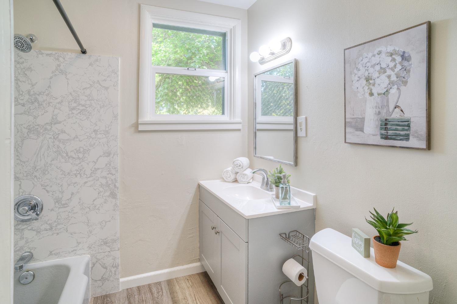 Bathroom with a gray vanity, window, and bathtub with a marble-patterned surround.