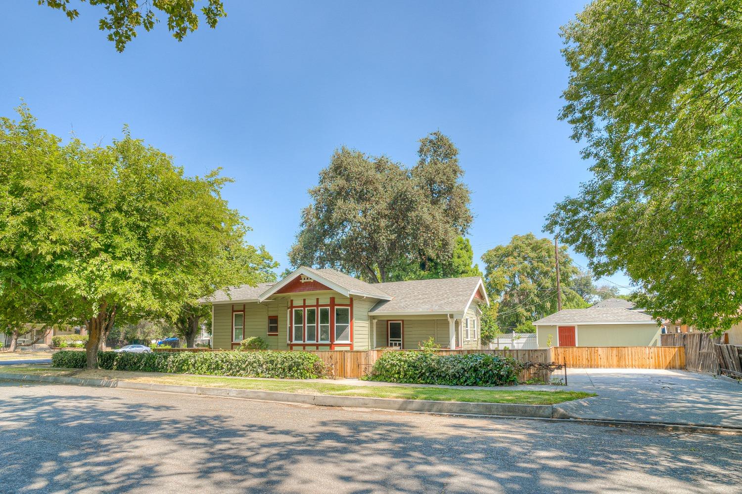 Sage-green single-story house with red trim, a wooden fence, and mature trees.