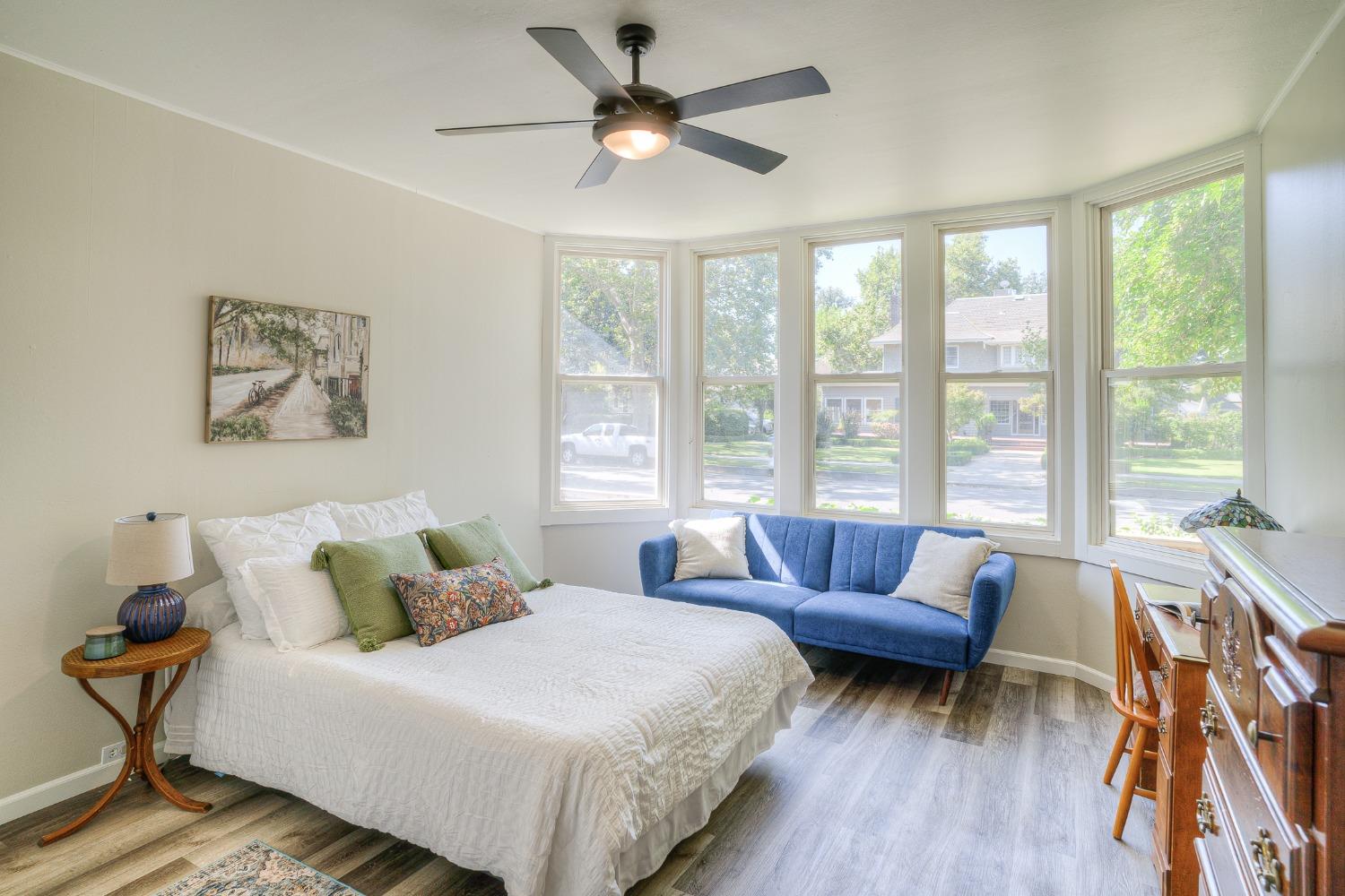 Bedroom with a white bed, blue loveseat, and large windows overlooking a tree-lined street.