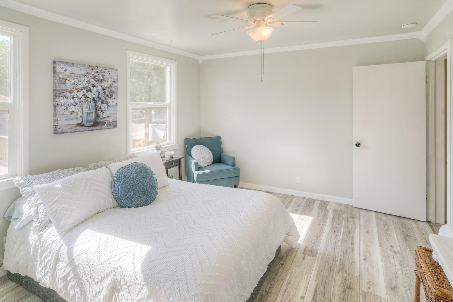 Bright bedroom with a white-covered bed, teal accent chair, and light wood-look flooring.