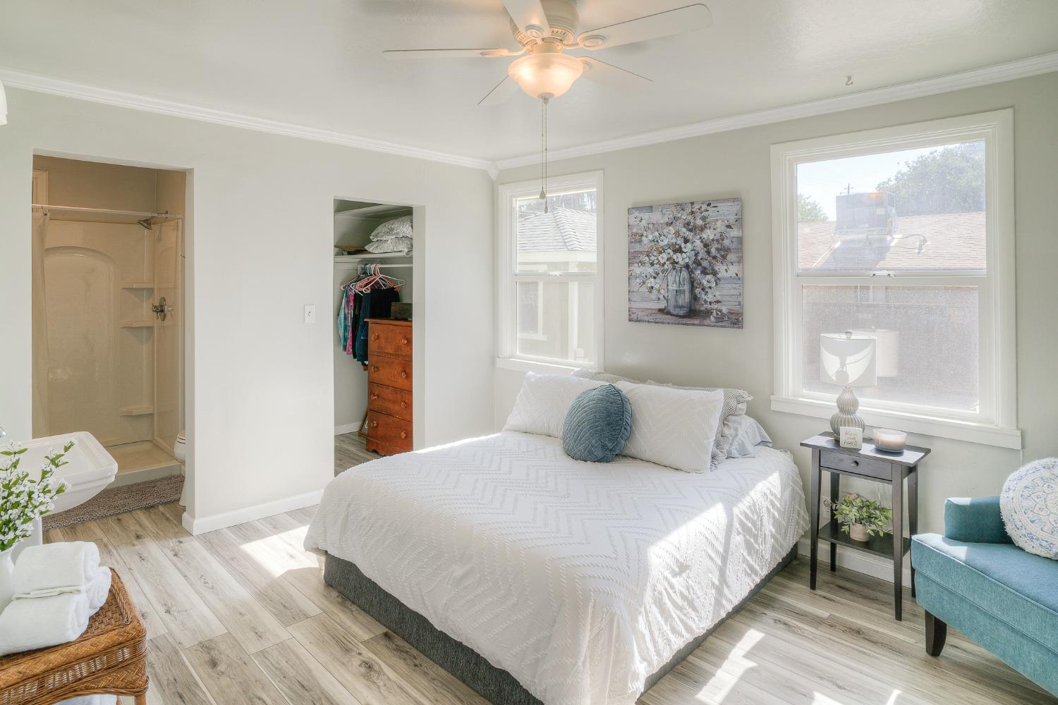Bright bedroom with a white bed, teal chair, ceiling fan, and adjoining shower.