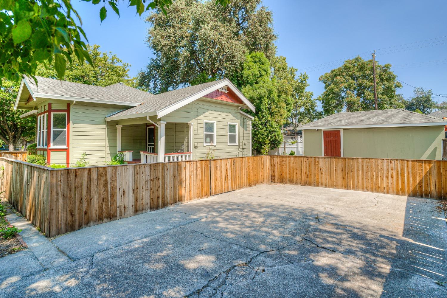 Sage-green house with red trim and covered porch, wooden fence, detached garage, and concrete driveway.