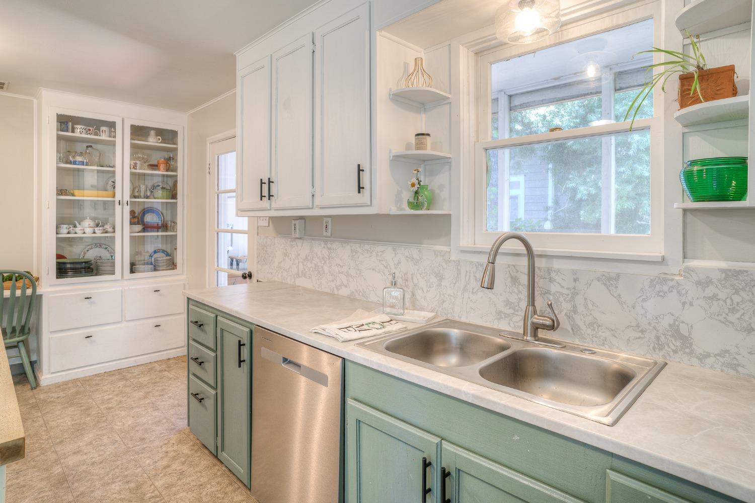 Kitchen with pale green cabinets, marble-patterned backsplash, and double-basin sink beneath a window.
