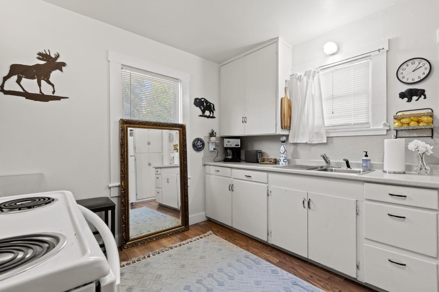 White kitchen with a coil stove, long counter, sink beneath a window, and a large leaning mirror.