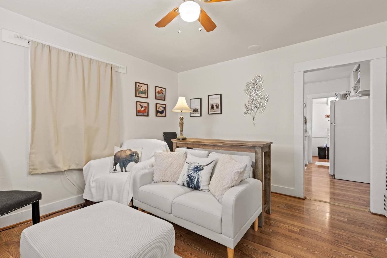 Living room with white sofas, cream curtains, and hardwood floors beside an open kitchen doorway.