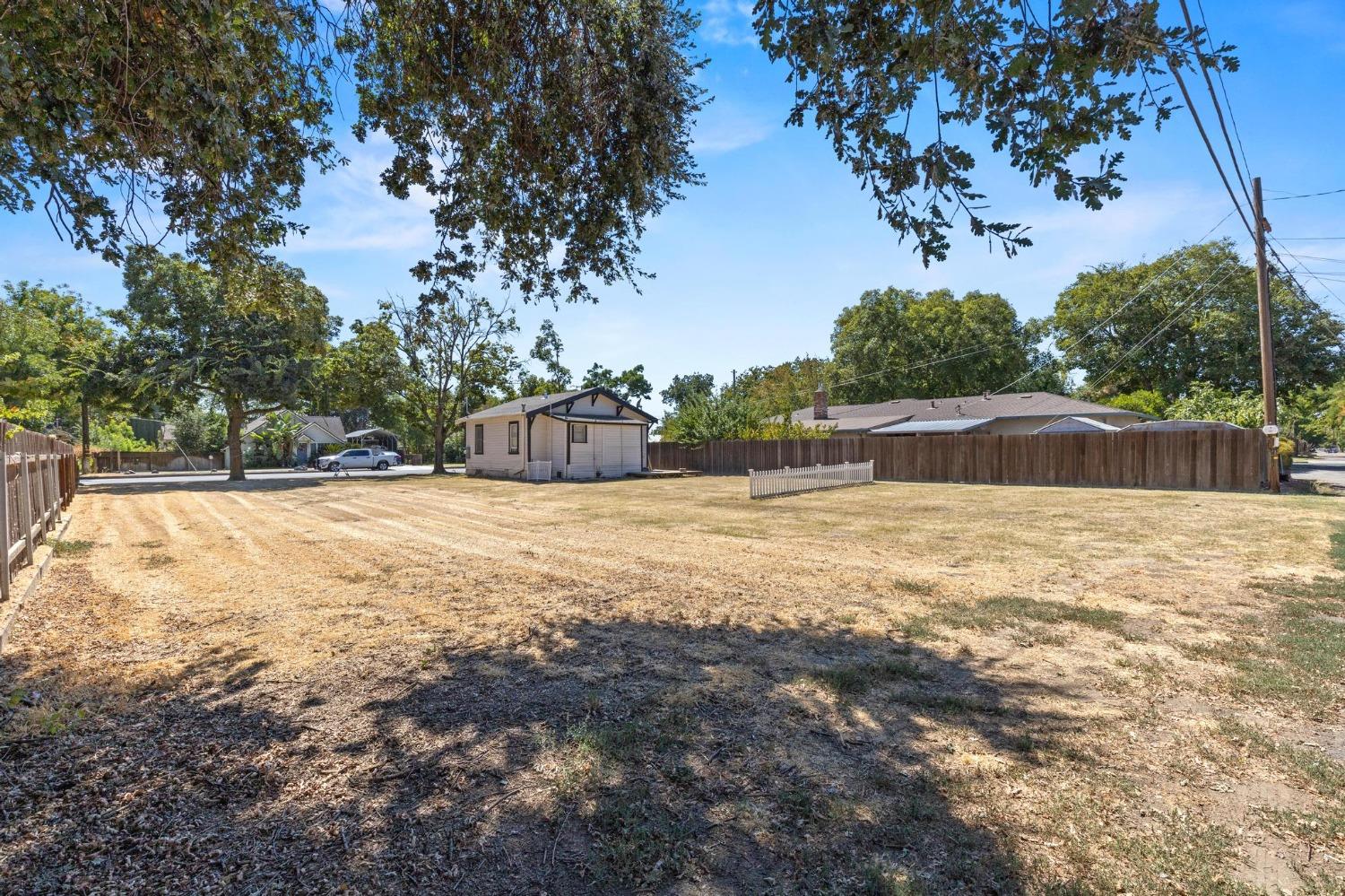 Small single-story house behind a broad dry yard, shaded by mature trees and bordered by wooden fencing.