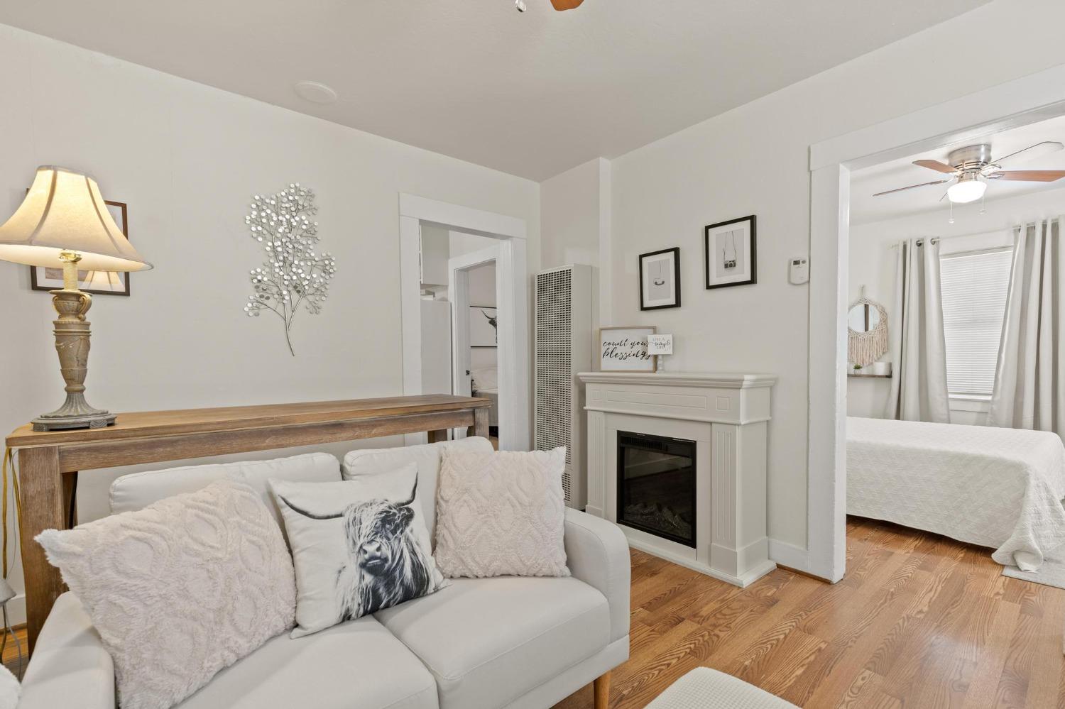 Living room with a white sofa, decorative fireplace, hardwood floors, and an adjoining bedroom.