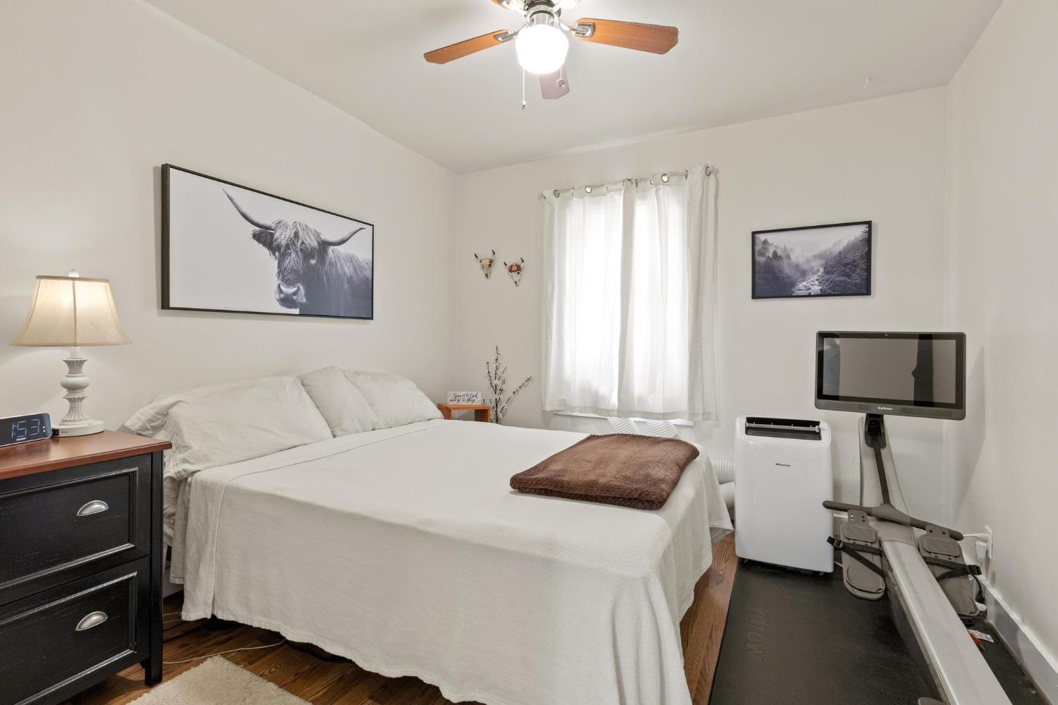 Bedroom with a white-covered bed, hardwood floor, ceiling fan, and exercise equipment.