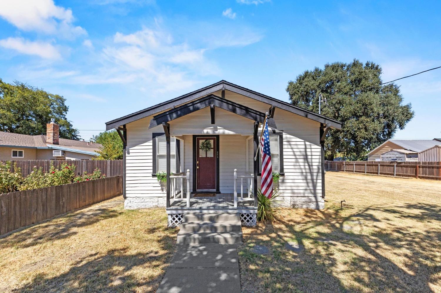 Gray single-story house with a covered porch, American flag, and fenced dry yard.