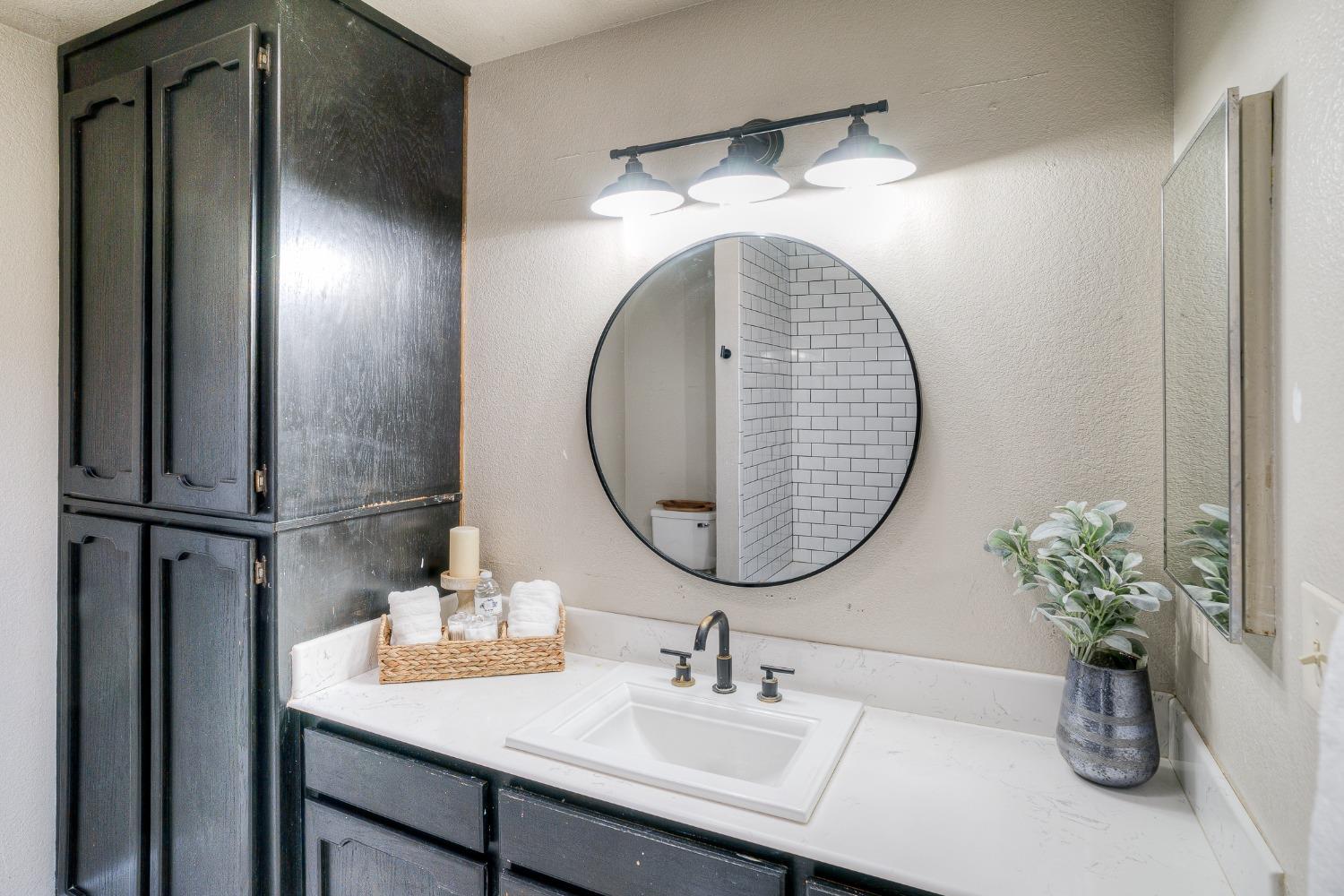 Bathroom vanity with dark cabinetry, white countertop, round mirror, and reflected subway-tile shower.