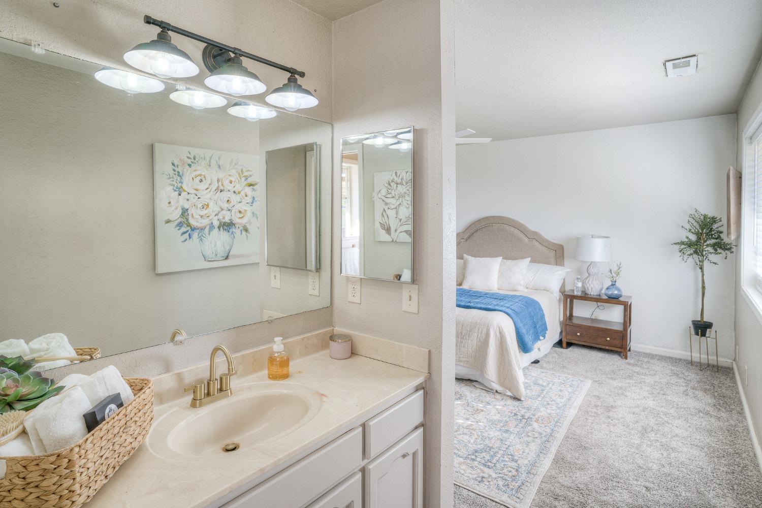 Bathroom vanity with a large mirror overlooking a bedroom with a white bed and blue throw.