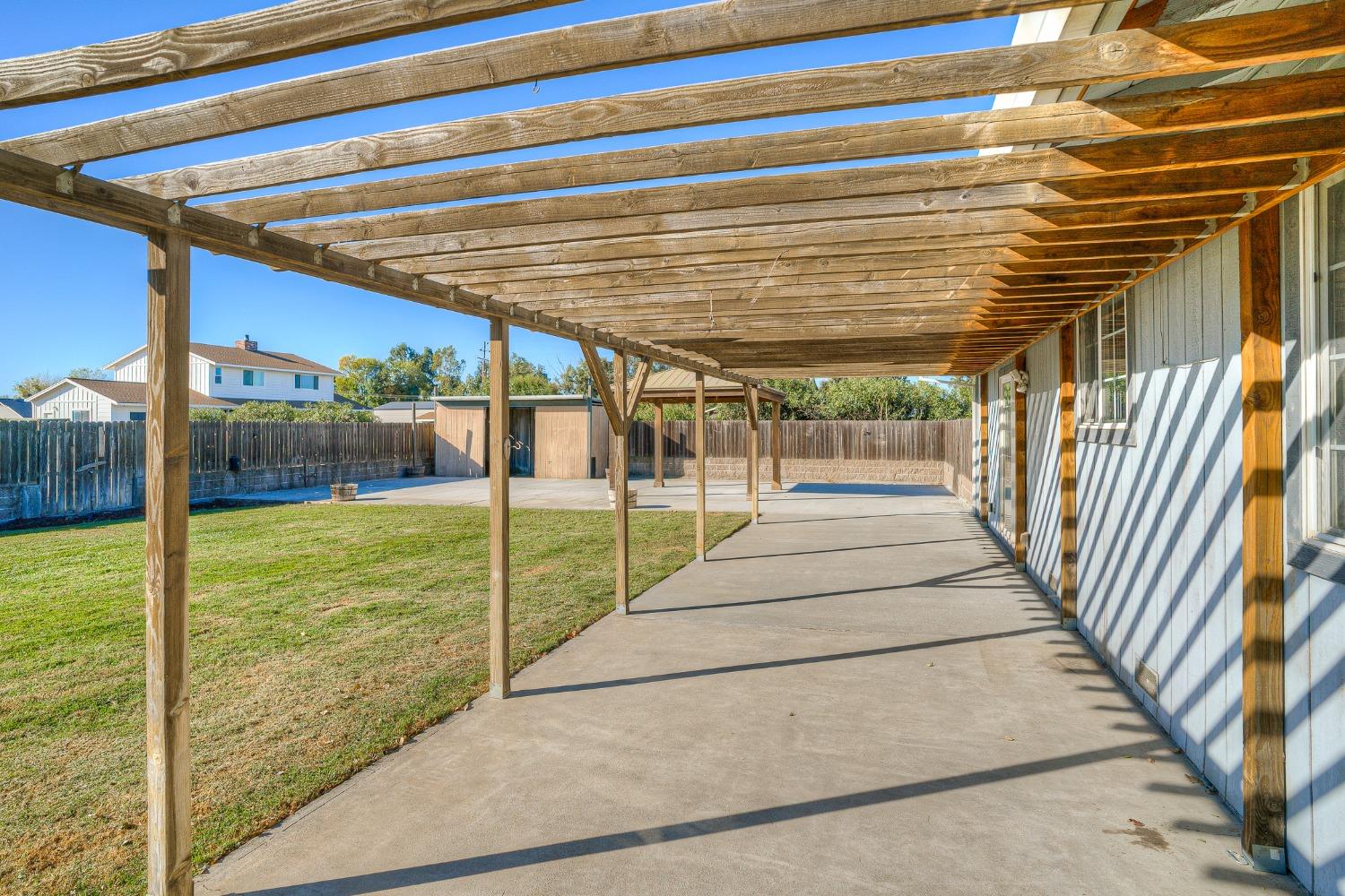 Backyard patio beneath a long wooden pergola, with lawn and a fenced yard.