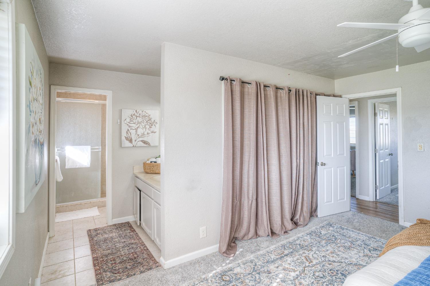 Bedroom with mauve curtains and patterned rugs, opening to a bathroom with a vanity and glass shower.